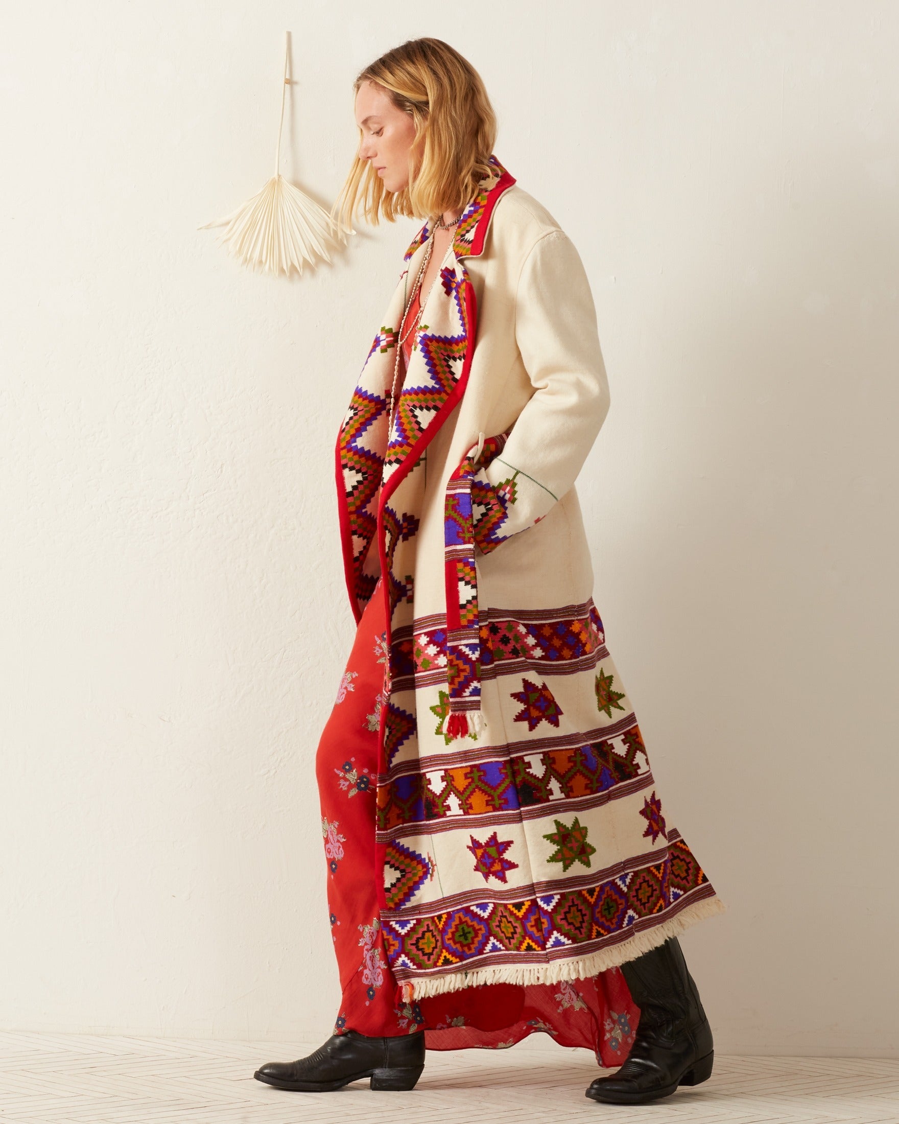 A woman in a colorful Penelope Tiger Eye Blanket Trench and red dress walks along a neutral wall. Her black boots complement the trench coat's intricate Southwest-style geometric patterns. A decorative hanging adds charm to the scene, enhancing the vibrant visual tapestry.