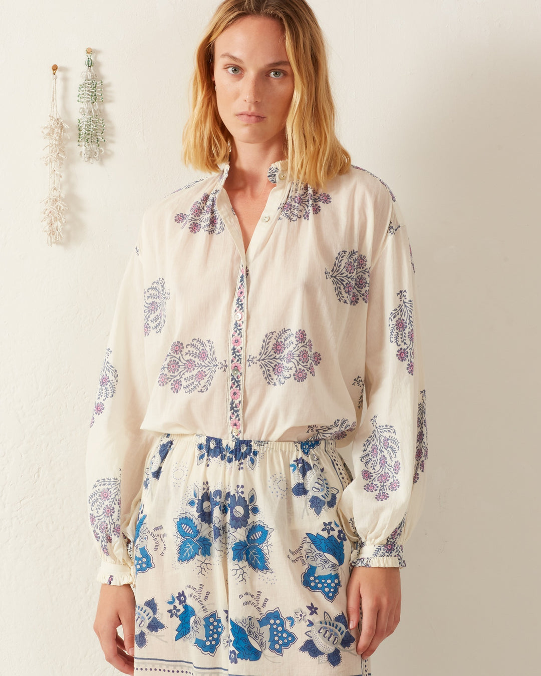 A person wearing the Poet Freesia Phlox Blouse, a white, long-sleeved top adorned with a blue and purple floral print, stands against a plain background. The button-up front and gathered waist evoke charm and allure. Dried plants hanging on the wall behind them enhance the vintage-inspired folk blouse vibe of the scene.