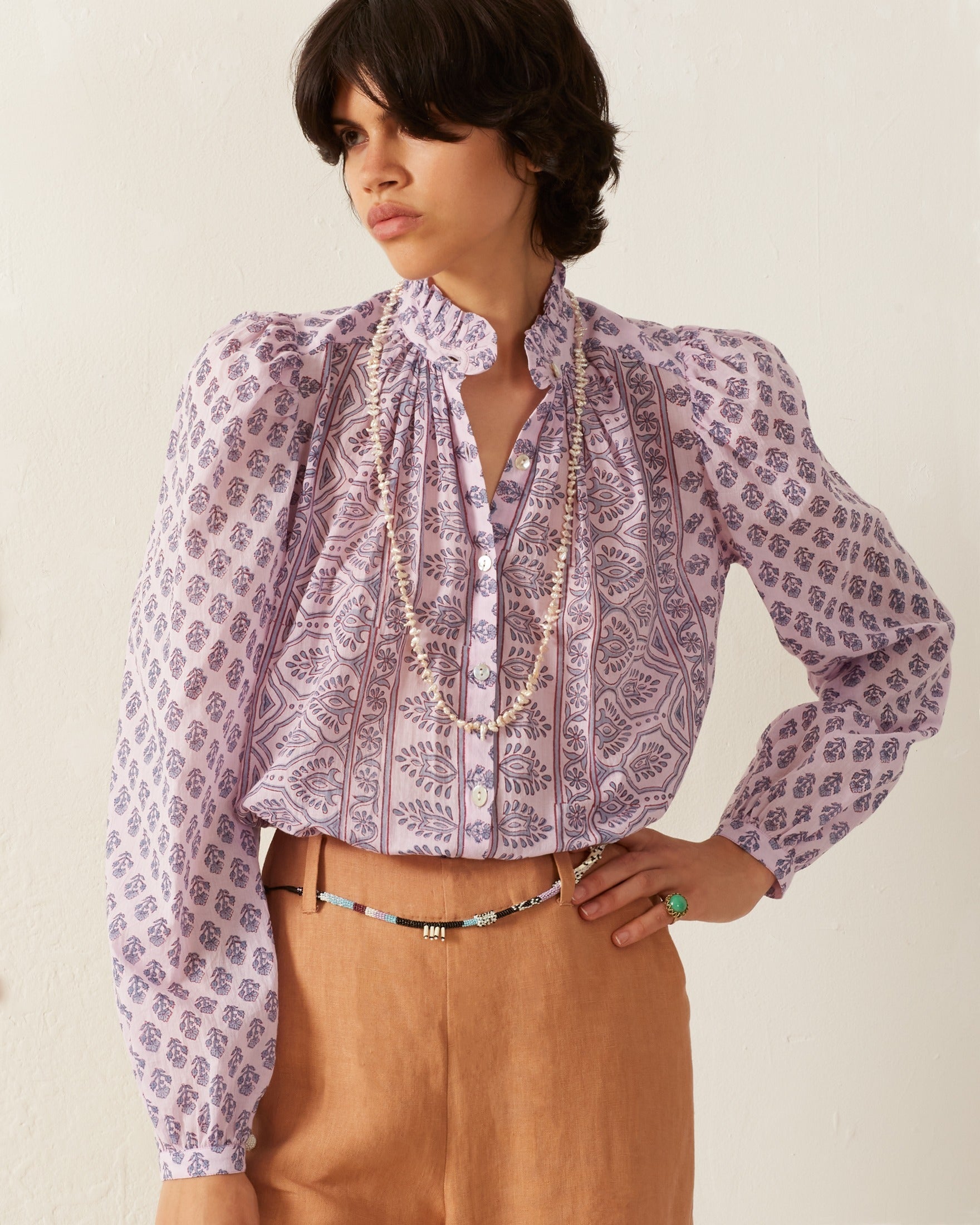 A person stands against a plain background wearing the Annabel Lilac Bhutti Shirt, which features a vintage-inspired purple pattern with ruffles, paired with beige trousers. They are accessorized with a necklace and a beaded belt, and their hand rests on their waist.