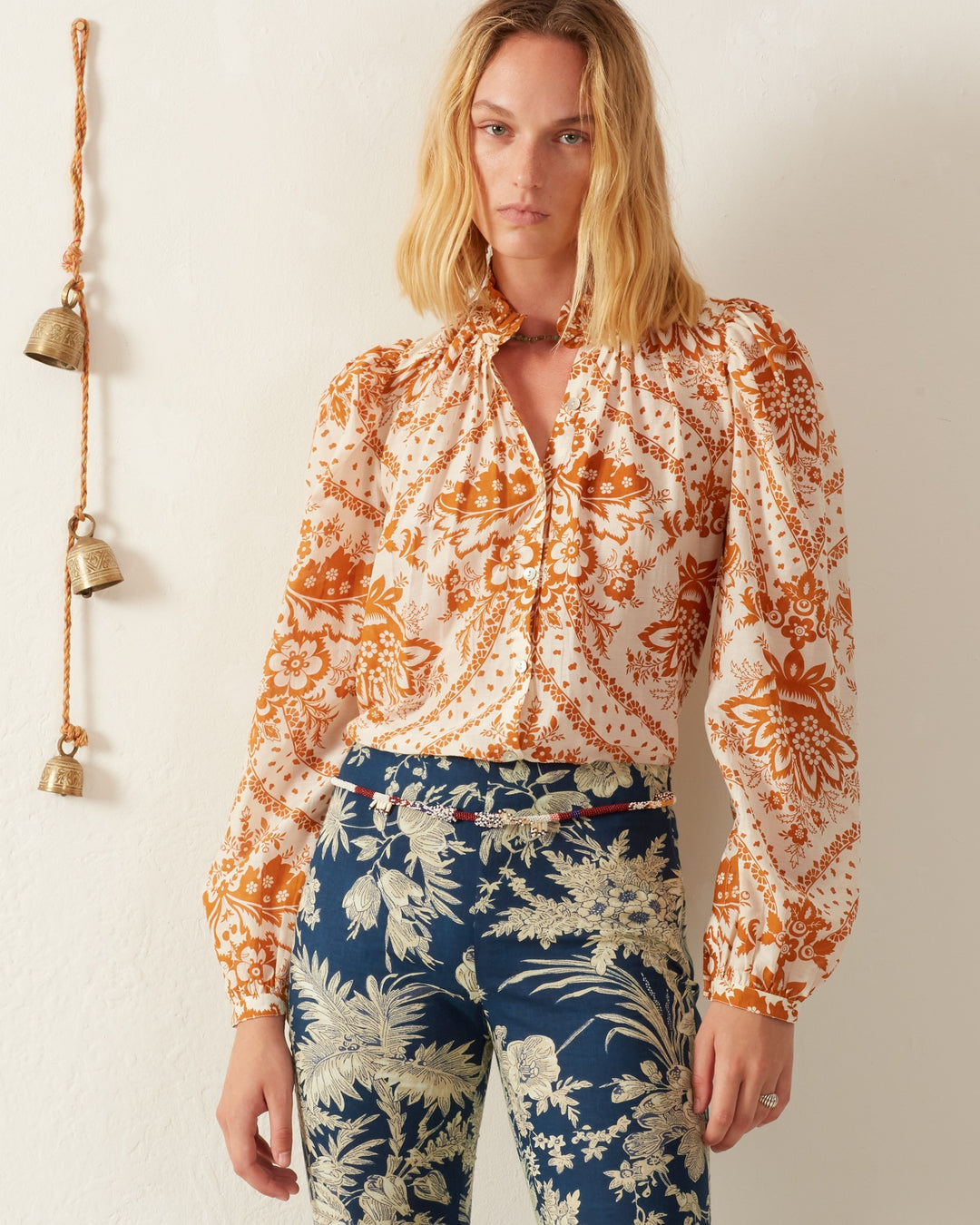 A woman with shoulder-length blonde hair is wearing the Annabel Sunglow Shirt, featuring a vintage-inspired orange floral motif, and pairs it with blue patterned pants. She stands against a light-colored wall adorned with decorative hanging bells.