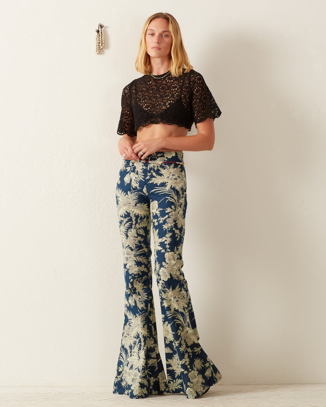 A person stands against a plain wall wearing the Nala Onyx Lace Cropped Top paired with blue high-waisted, wide-leg pants featuring a daisy motif. They have long, straight hair and are looking at the camera, with a small wall decoration hanging nearby.