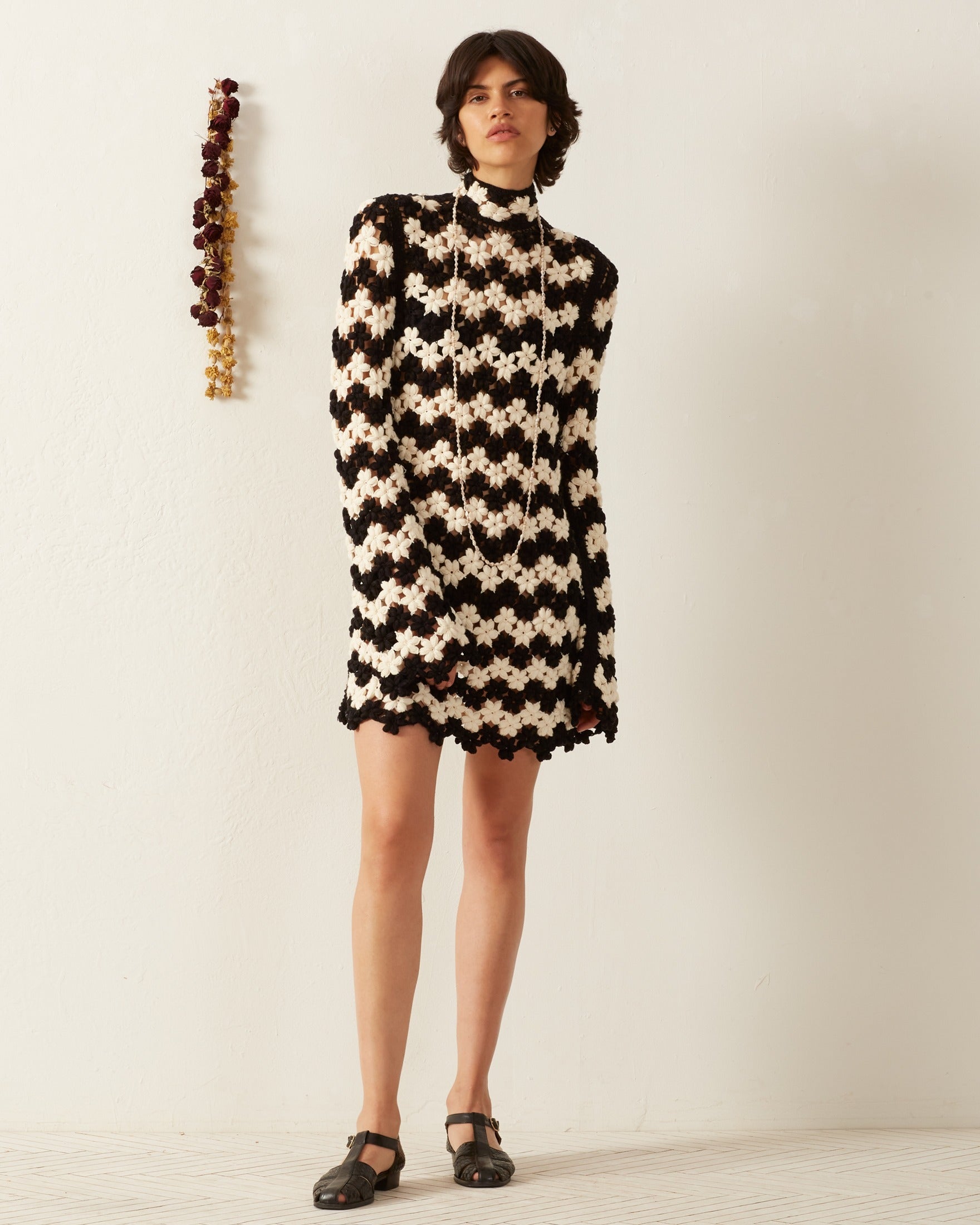 A person stands against a light-colored wall, wearing the Romy Mariner Crochet Dress and black shoes. A decorative string of dried flowers, adding a flower child touch, hangs on the wall beside them.