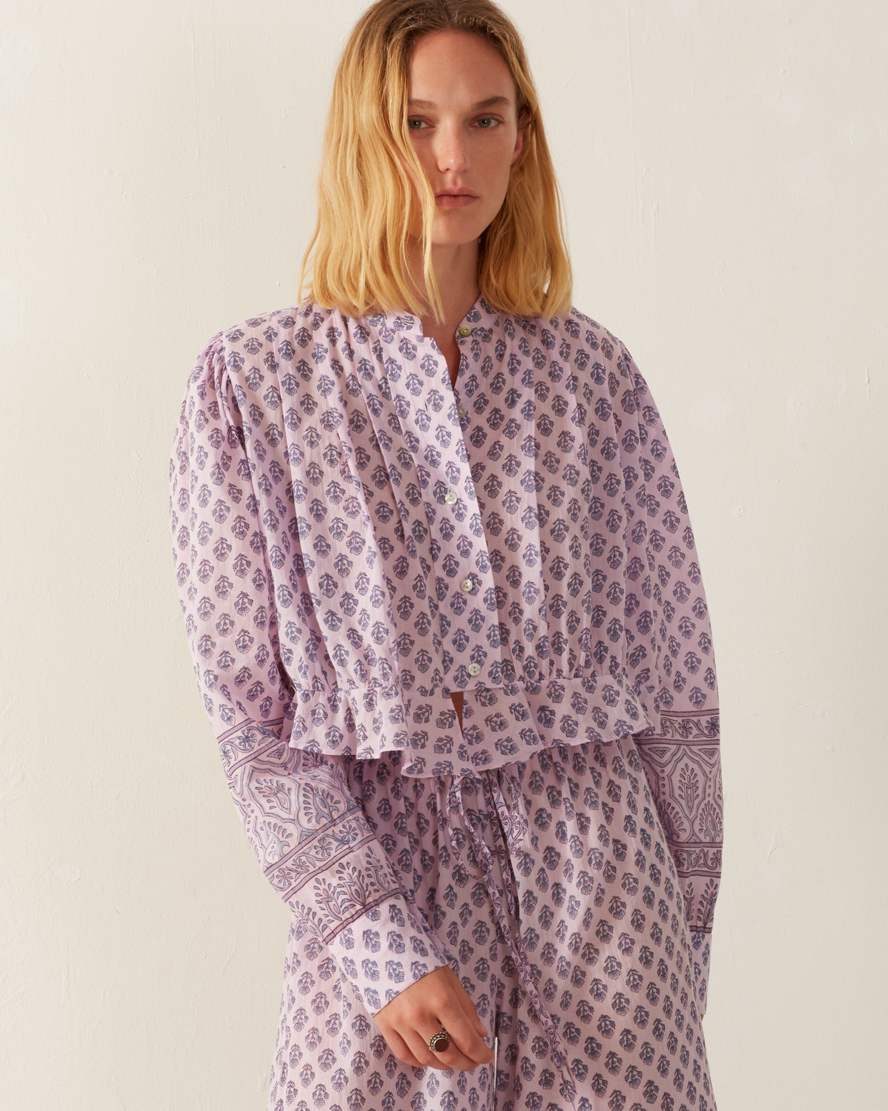 A person with long blonde hair is wearing the Ryland Lilac Bhutti Top, a lavender floral print blouse crafted from cotton voile, paired with matching pants. The outfit showcases intricate designs reminiscent of a Victorian silhouette against a plain, light-colored wall.