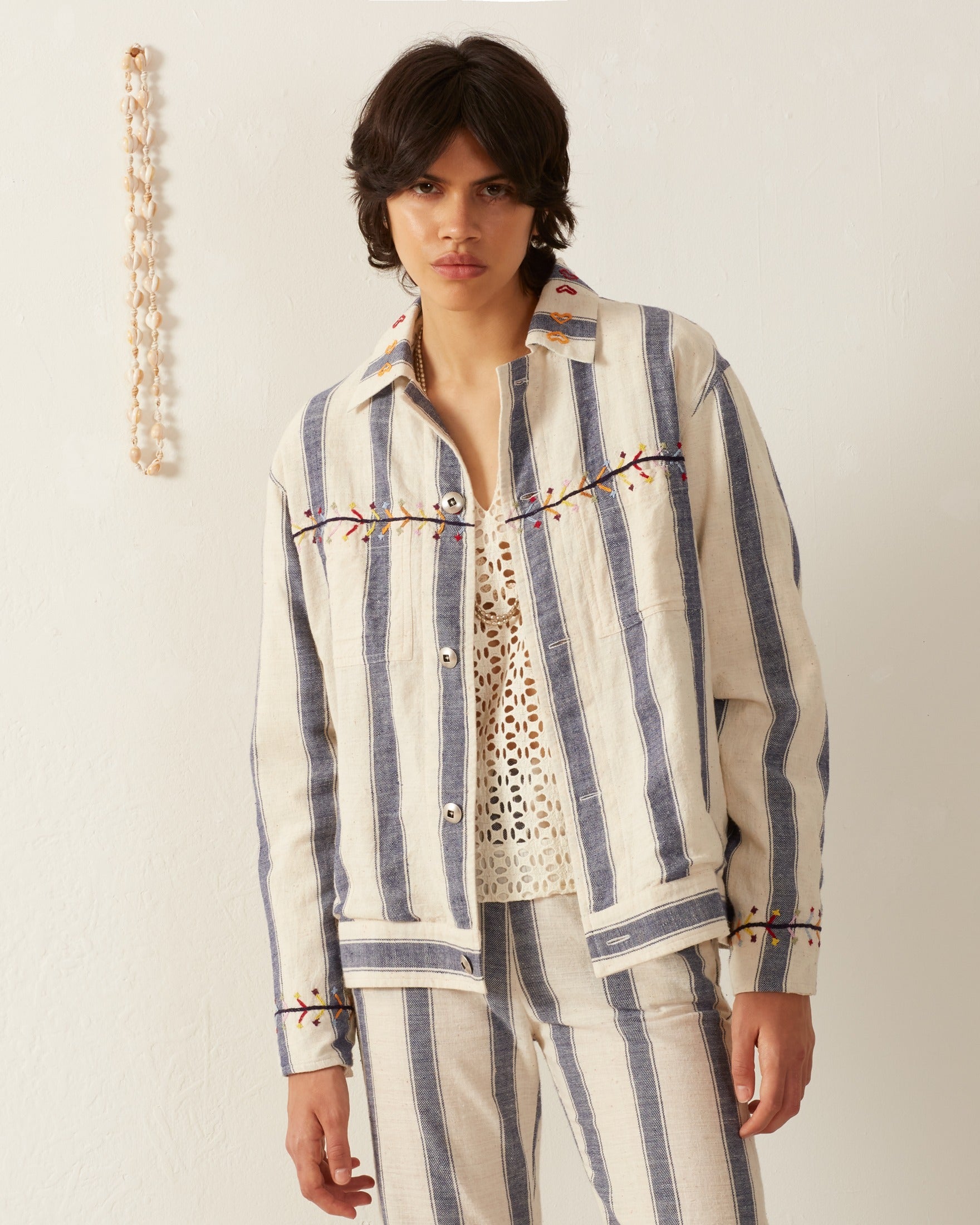 A person wearing the Seba Arrow Stripe Jacket and matching pants set with hand-embroidered details stands against a plain white background. They are also wearing a lace-patterned top underneath the jacket. A decorative string hangs on the wall beside them.