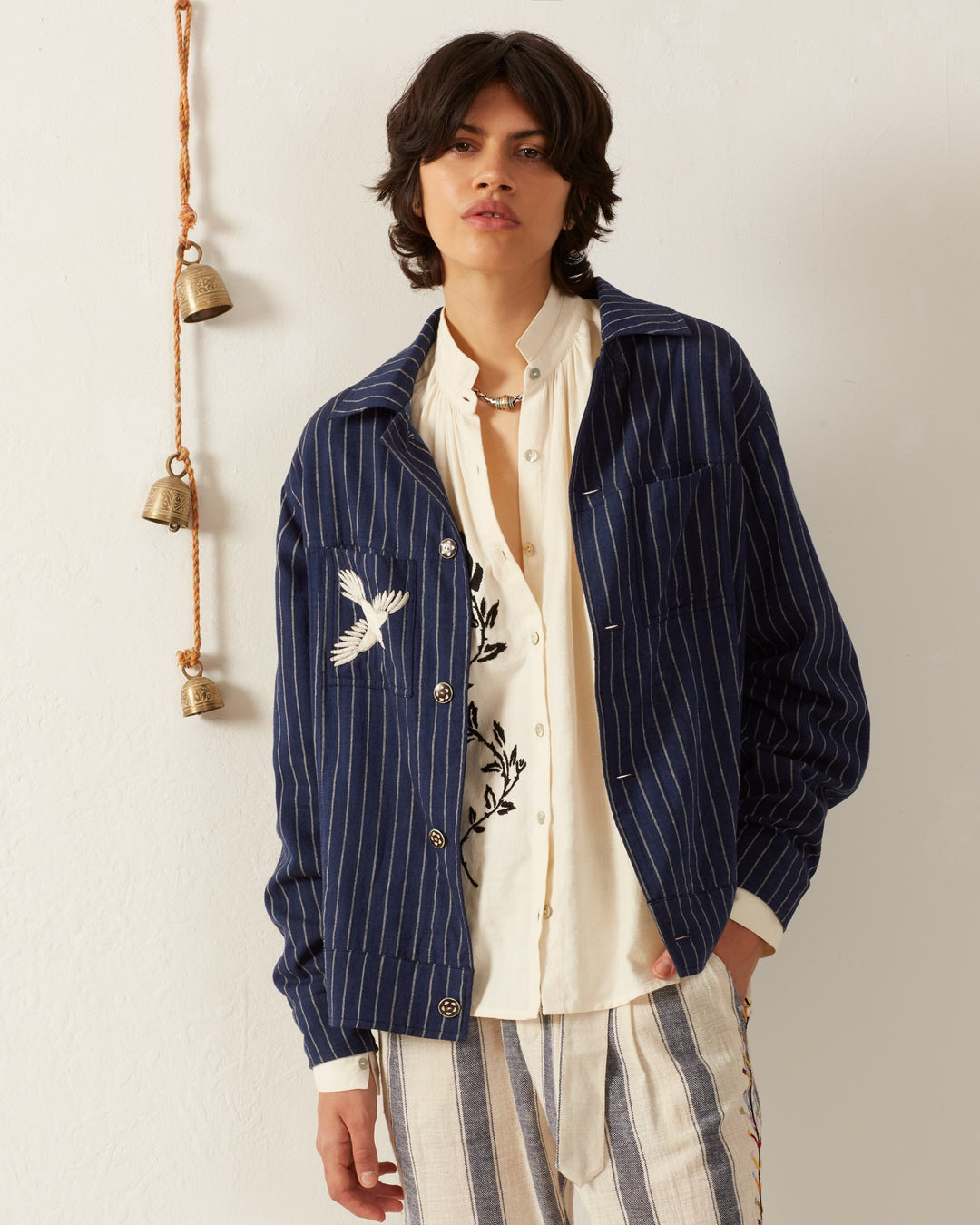 A person with medium-length dark hair wears the Seba Luna Night Jacket, showcasing a navy striped design with a bird print, over a cream shirt. They stand against a white wall decorated with hanging brass bells, paired with light striped pants crafted from hand-embroidered Khadi cotton.