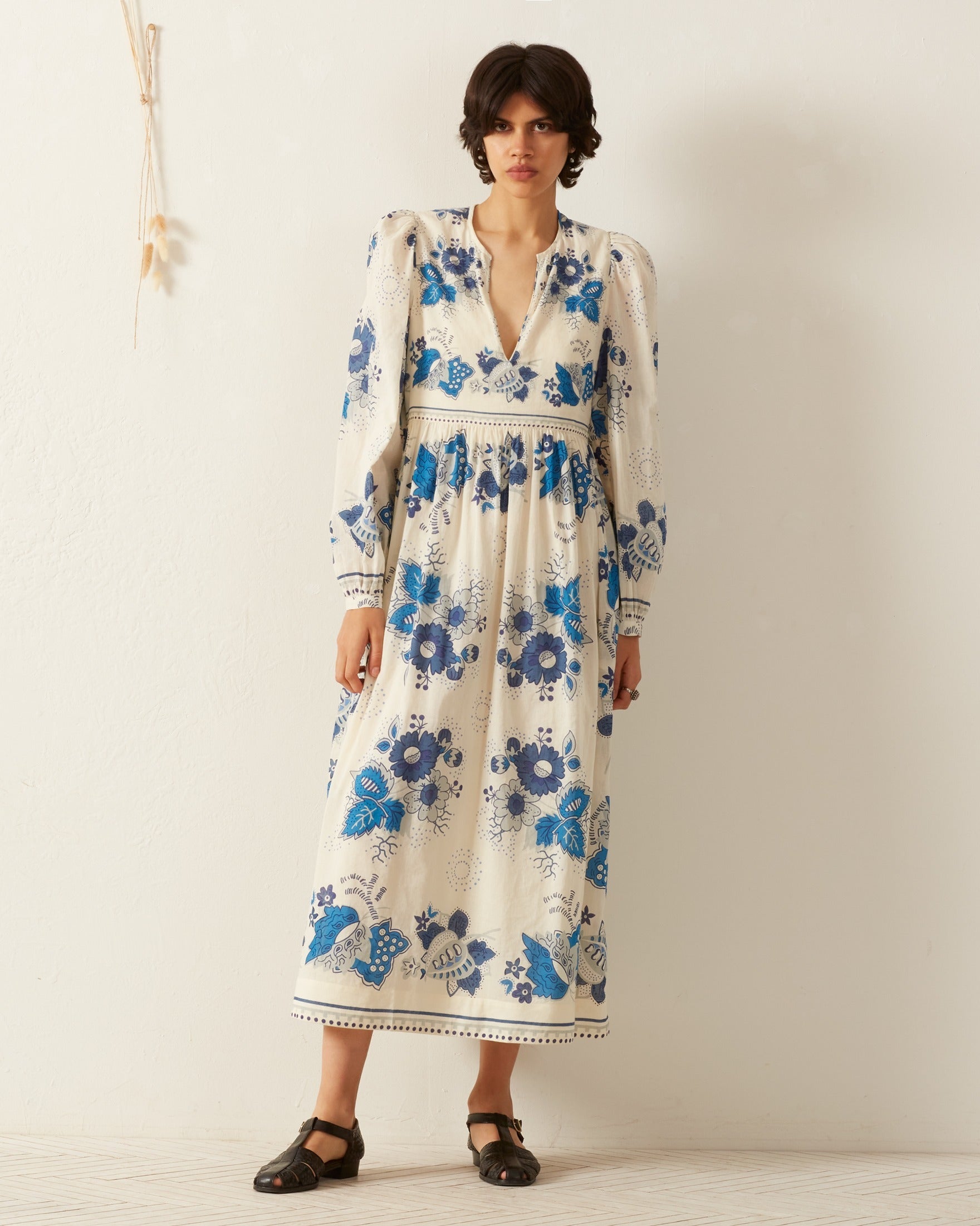 Dressed in the Winifred Blue China Panel Dress, which features a blue-and-white floral pattern, a person stands in a minimalist setting. The cream-colored dress is designed with puffed long sleeves and a deep V-neck. Their ensemble is complemented by black open-toed sandals, with dried flowers artfully hanging on the wall behind them.