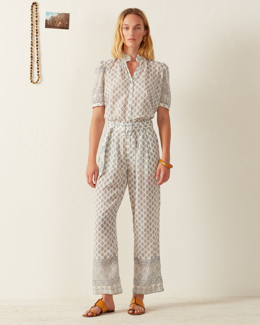 A woman poses against a plain wall, wearing the Winnie Delft Bhutti Shirt, a vintage-inspired cotton voile short-sleeve blouse paired with matching pants. She completes her look with a yellow bracelet and brown sandals, while a beaded necklace and photo adorn the wall behind her.