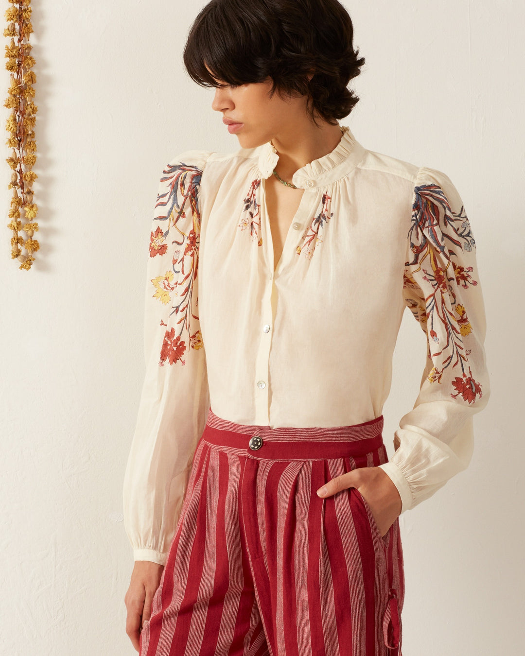 A person with short dark hair is wearing the Annabel Sun Lily Valley Shirt, a cream blouse featuring hand block-printed floral motifs on the shoulders and long sleeves. Complementing it are high-waisted red and white striped pants. They stand against a plain light-colored wall adorned with dried flowers, which enhances the elegance of the cotton voile fabric.