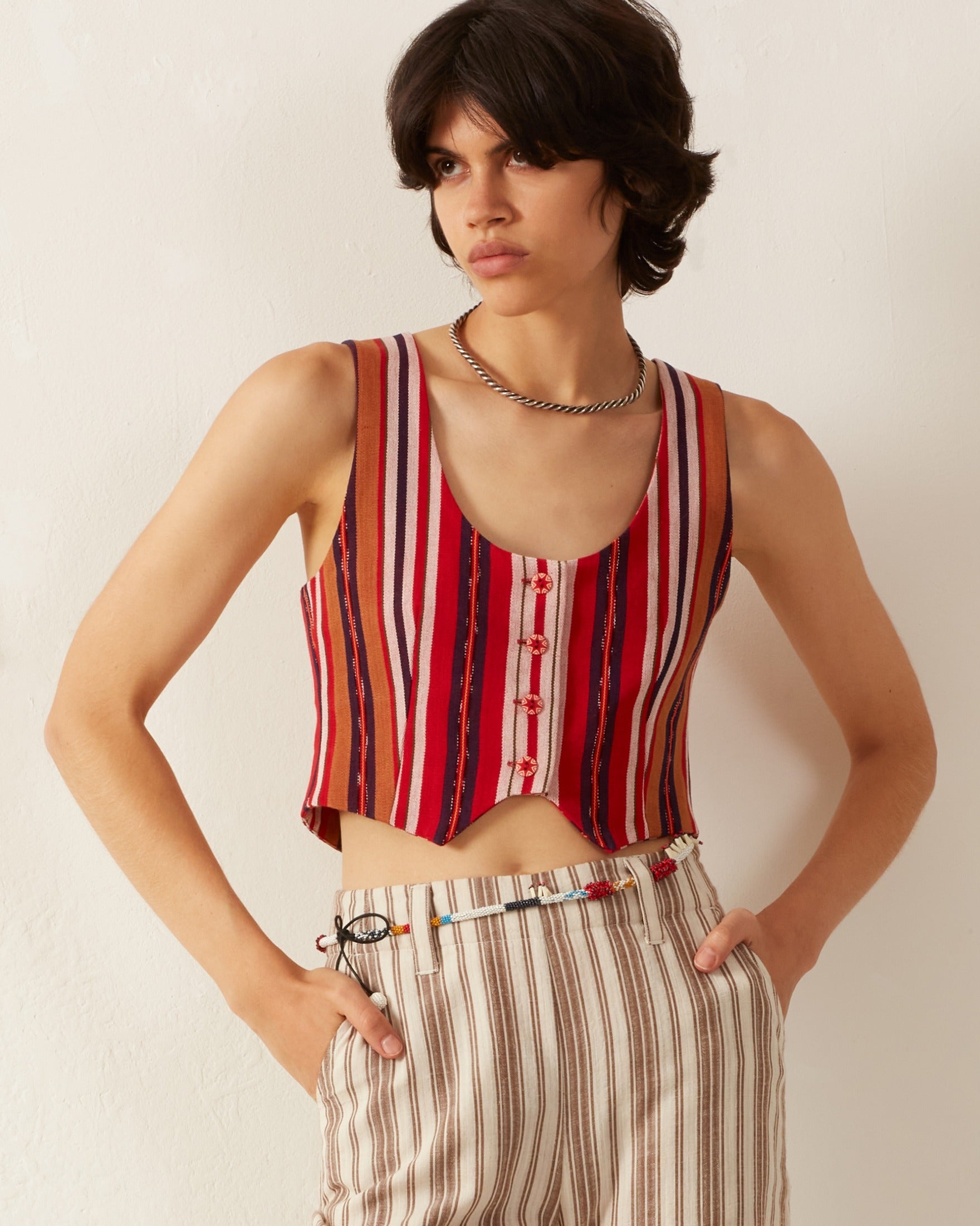 A person with short, dark hair wears the sleeveless, fitted Mossie Canyon Stripe Top in red and white with cloth-covered buttons, paired with high-waisted striped pants. They pose against a plain white background with one hand on their hip, adorned with a silver chain necklace.