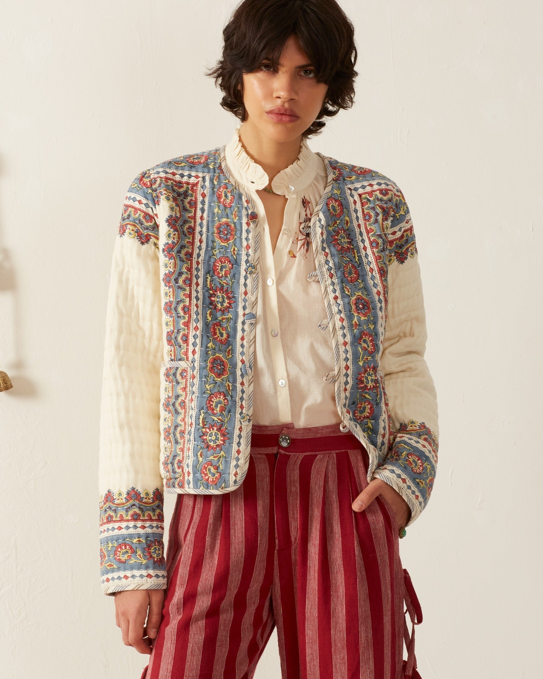 A person with short dark hair wears the Pasha Riviera Jacket, showcasing its detailed block-print motif, over a white blouse and red striped pants, standing against a plain backdrop.