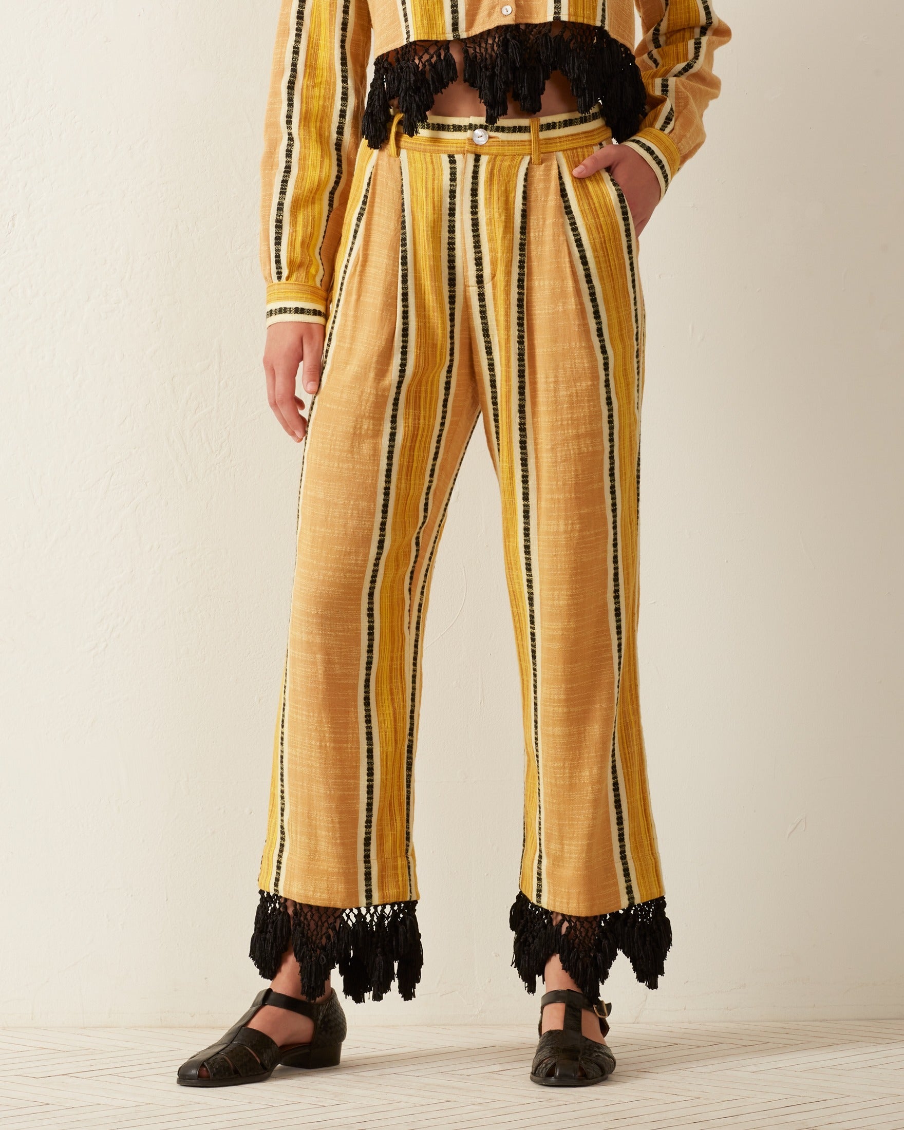 A person is wearing the Colette Sun Stripe Fringe Pant in mustard yellow, showcasing stripes and black fringes at the hem. Made from hand-loomed cotton, these high-waisted pants are paired with a matching top, beautifully highlighting the vertical stripes and black accents. The ensemble is completed with black sandals for a chic appearance.