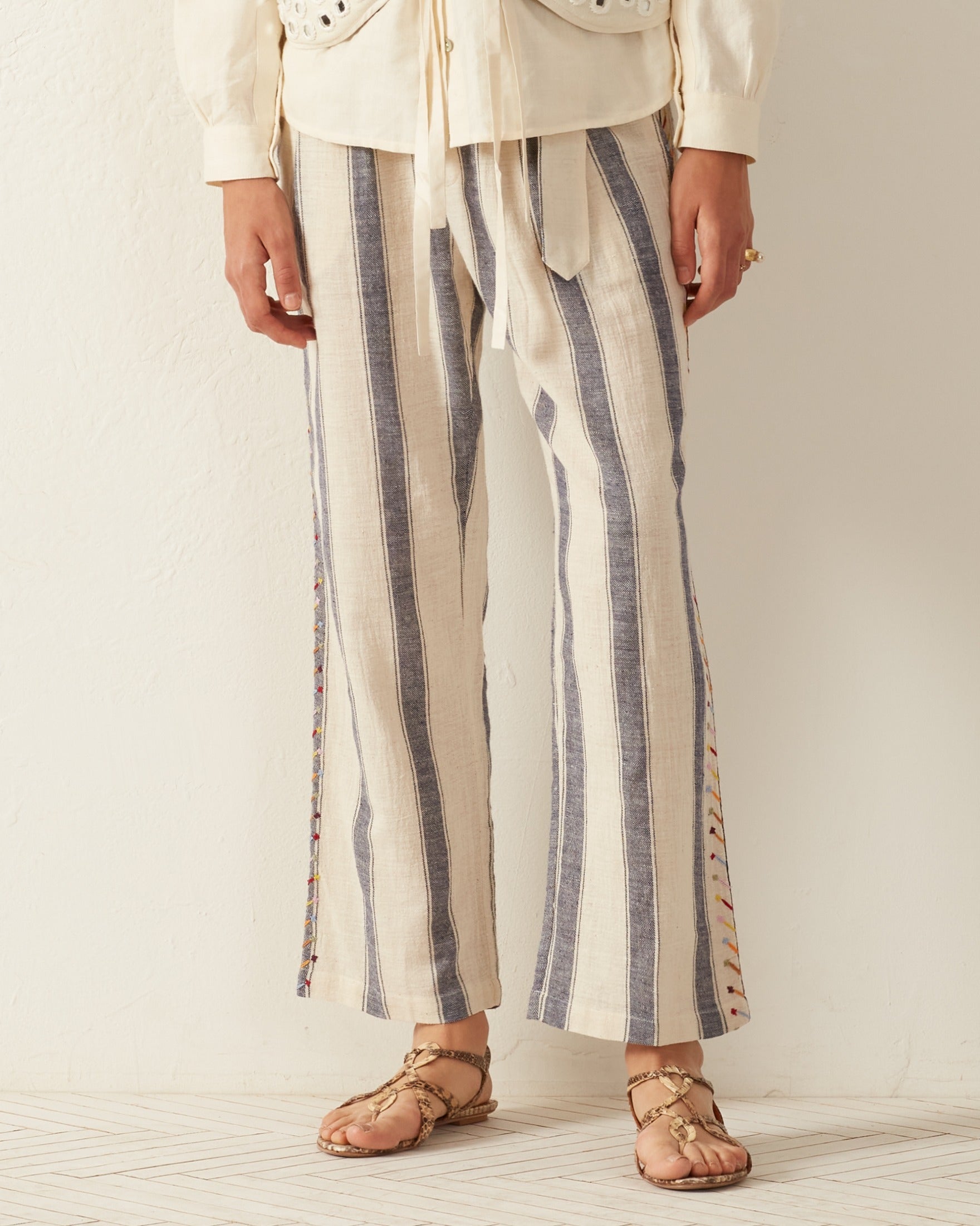 A person dressed in the Colette Arrow Stripe Pant, featuring cream and blue vertical stripes and a high-waisted design, stands on a light wooden floor wearing beige sandals. The pants are made from hand-loomed cotton with a relaxed fit and decorative side details against a plain cream wall backdrop.