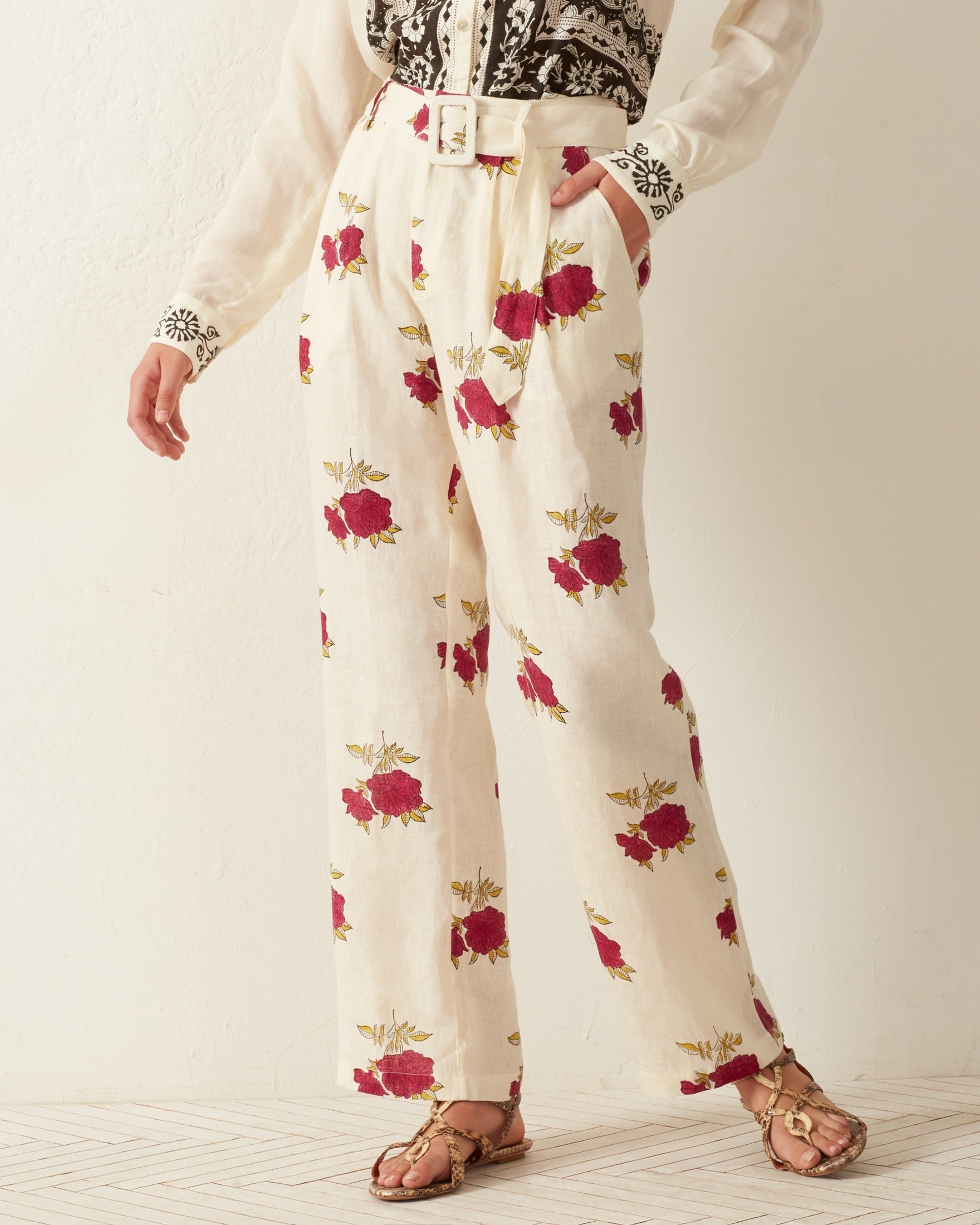 A person is wearing the Colette Cerise Rosette Pant, featuring a cream color with a high waist and pink rose print, complete with a matching belt. They also have on a white blouse with black embroidered details and are standing on a wooden floor.