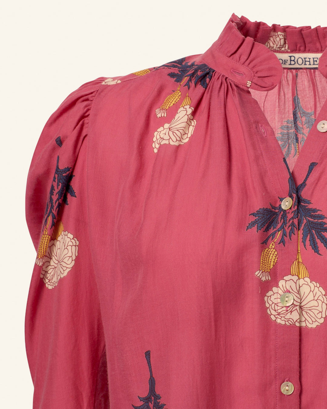 The Arte Hibiscus Rose Shirt showcases a vintage vibe with its close-up of pink fabric adorned with white and blue floral embroidery. This shirt features a high ruffled collar, button-down front, and intricate hand-embroidered details. The lightweight fabric is slightly gathered at the shoulders for an elegant finish.