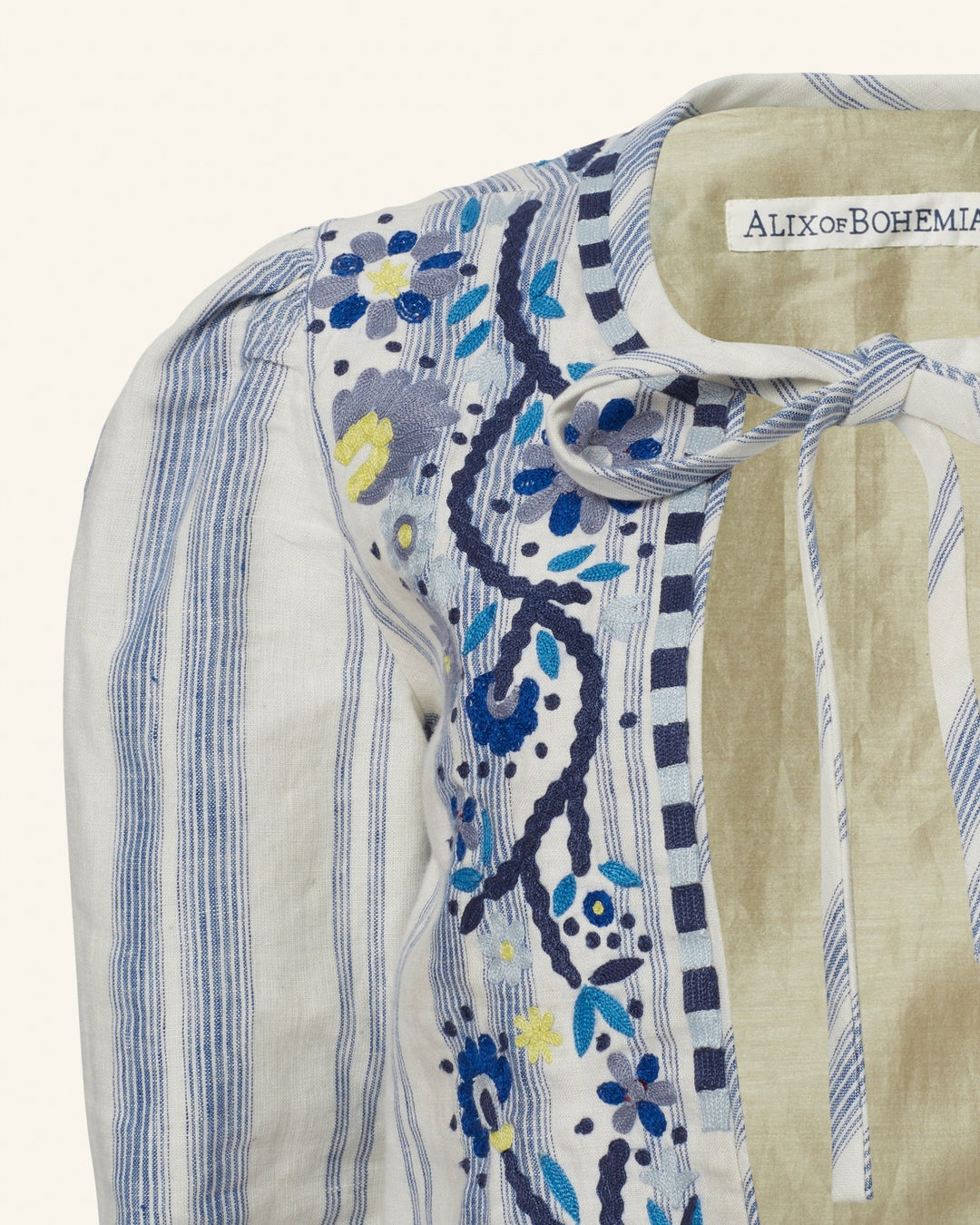 Close-up of a garment showcasing part of the Fiorello Pacific Reef Jacket with blue and white vertical stripes. The jacket, hand-embellished with intricate floral embroidery in shades of blue and yellow, features a label reading "Alix of Bohemia" and a tie detail at the collar.