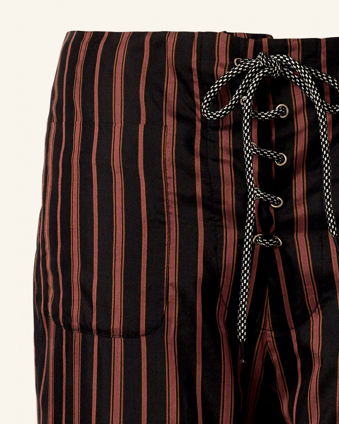 A close-up of the Jodphur Clove Beaded Pant reveals its hand-block printed linen design, showcasing black pants adorned with vertical red and brown stripes. The pants are equipped with a stylish black and white braided lace-up closure at the front and feature two convenient side pockets.