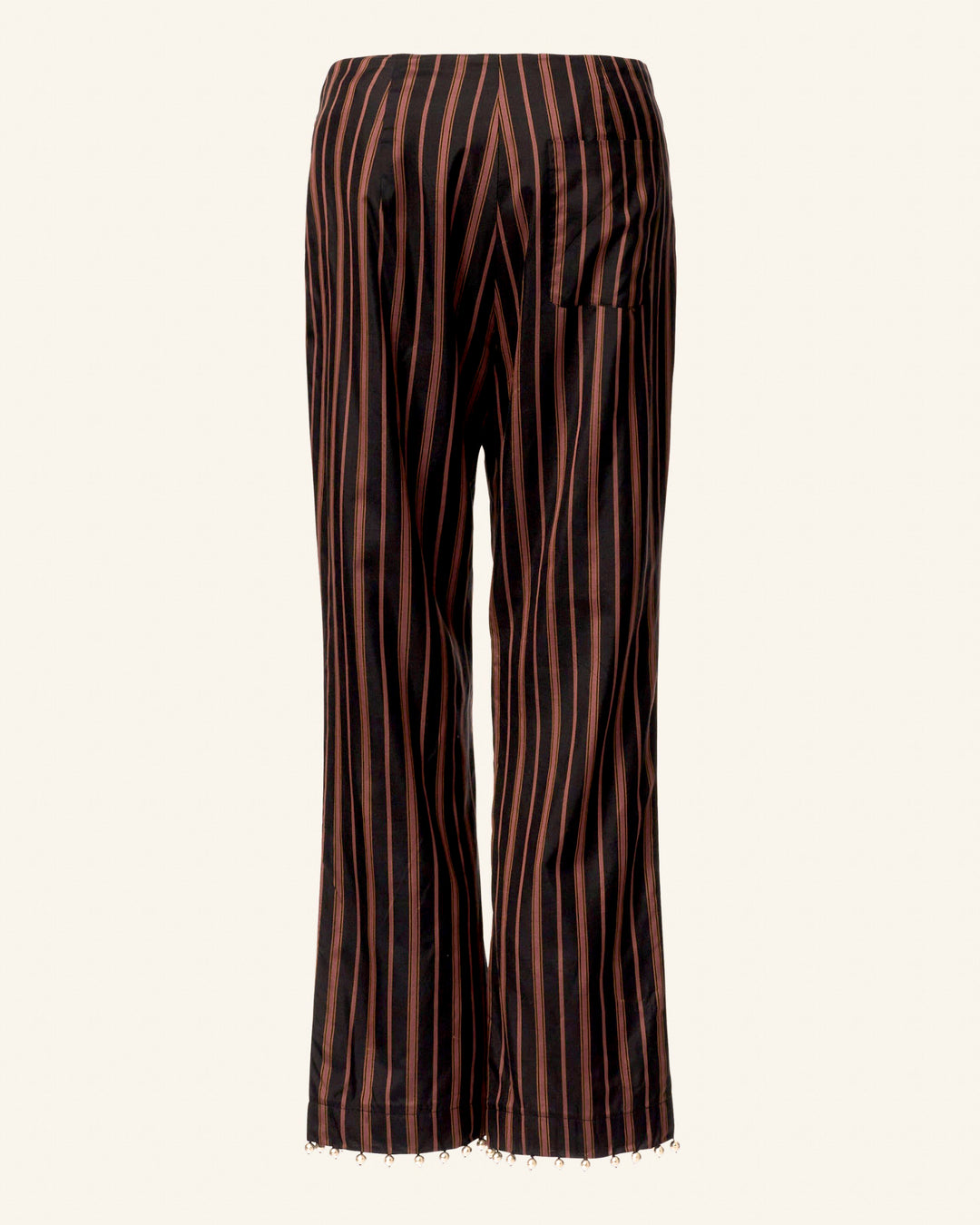 The Jodphur Clove Beaded Pant is a pair of black pants featuring vertical red stripes, a structured fit, and a waistband. These pants are accented with hand-block printed linen embellishments at the hem and include a single back pocket. They are presented against a plain white background.