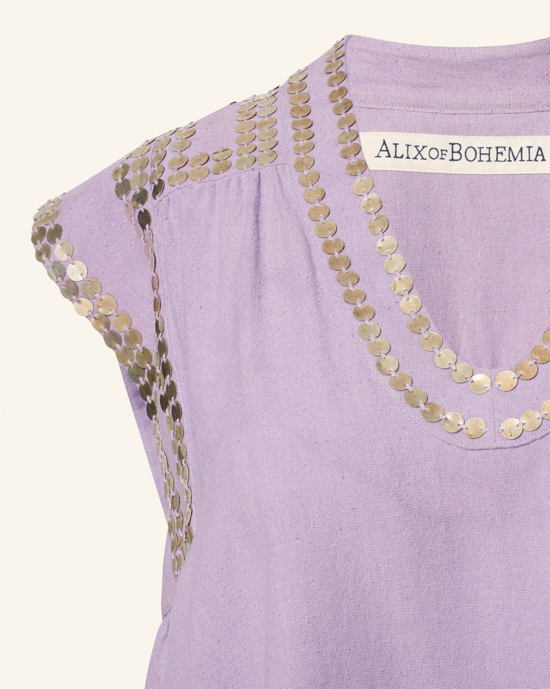 The Nehru Lavender Coin Top is a sleeveless Banaras silk piece in purple, adorned with gold sequin detailing around the neckline and shoulders. Labeled "Alix of Bohemia," it showcases exquisite hand-stitched silver coins, adding a unique touch to its elegance.