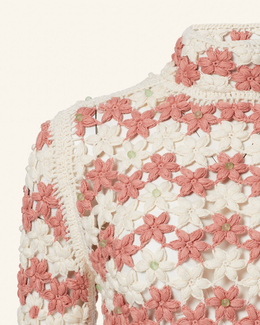 A detailed close-up of the Romy Peachy Crochet Dress highlights its khadi cotton fabric adorned with a floral pattern featuring pink and cream flowers. The dress's intricate design is complemented by small round green accents that add a delicate texture against a plain light backdrop. Pair it with high-waisted shorts for an effortlessly chic look.