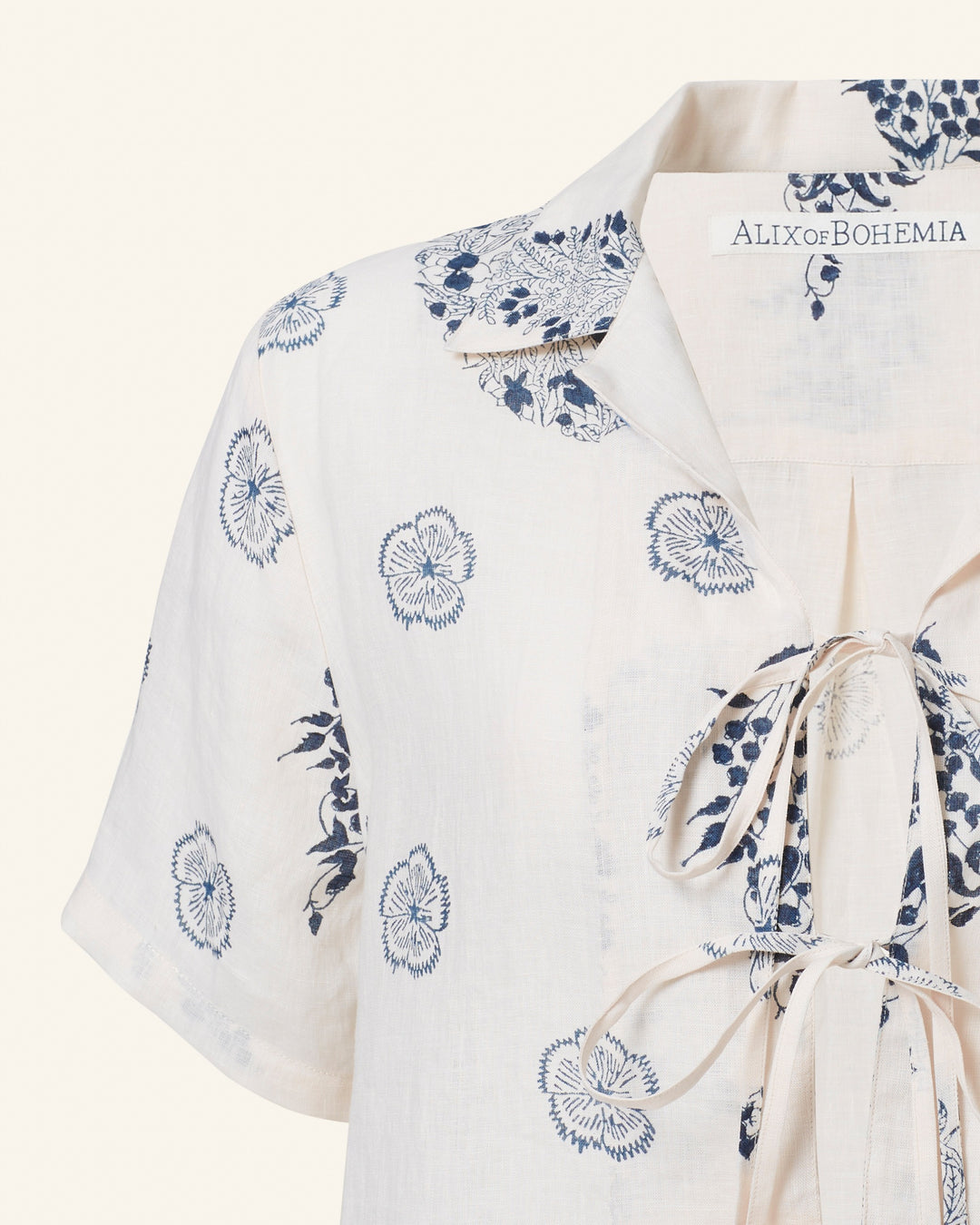 The "Styla Porcelain Linen Shirt" exudes a relaxed, bohemian charm with its white linen fabric, blue floral patterns, and lace-up detail on the front. Graced by the brand "Alix of Bohemia," it evokes a vintage Hawaiian shirt vibe. This short-sleeve piece offers effortless style with its laid-back flair.