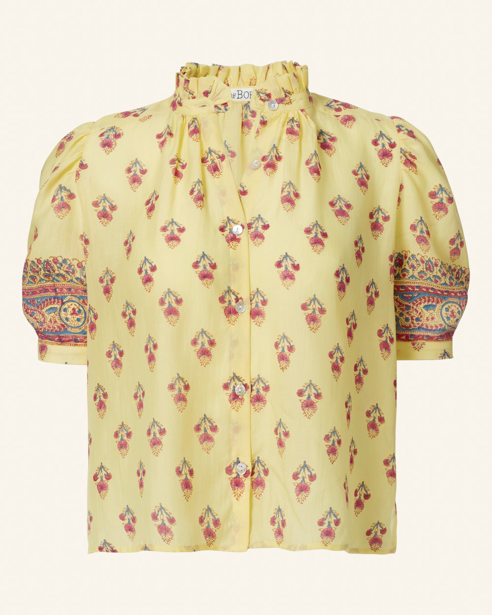 The Winn Primrose Bhutti Shirt is a vintage-inspired yellow blouse adorned with a pink and red floral pattern. It boasts short puffed sleeves highlighted by an intricate contrasting design, complemented by a ruffled high collar, all crafted from soft cotton voile. Elegant button closures run down the front.