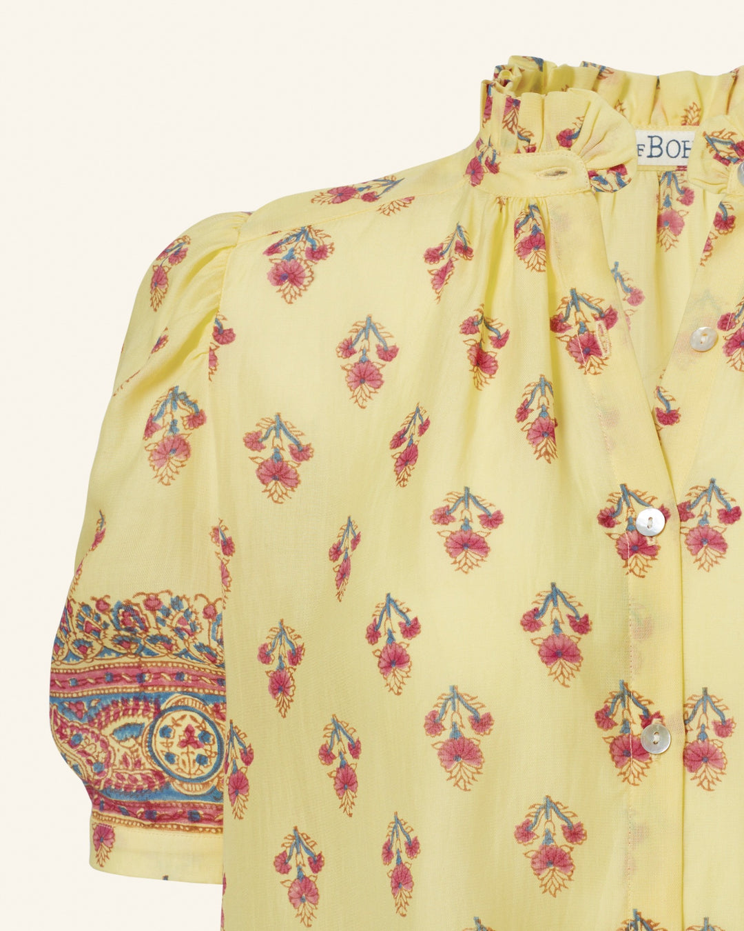 Close-up of the Winn Primrose Bhutti Shirt, a vintage-inspired yellow blouse featuring a floral motif with red and blue flowers. It boasts puffed sleeves adorned with detailed multicolored embroidery on the cuffs and a ruffled collar. This cotton voile blouse is designed with shiny buttons down the front.