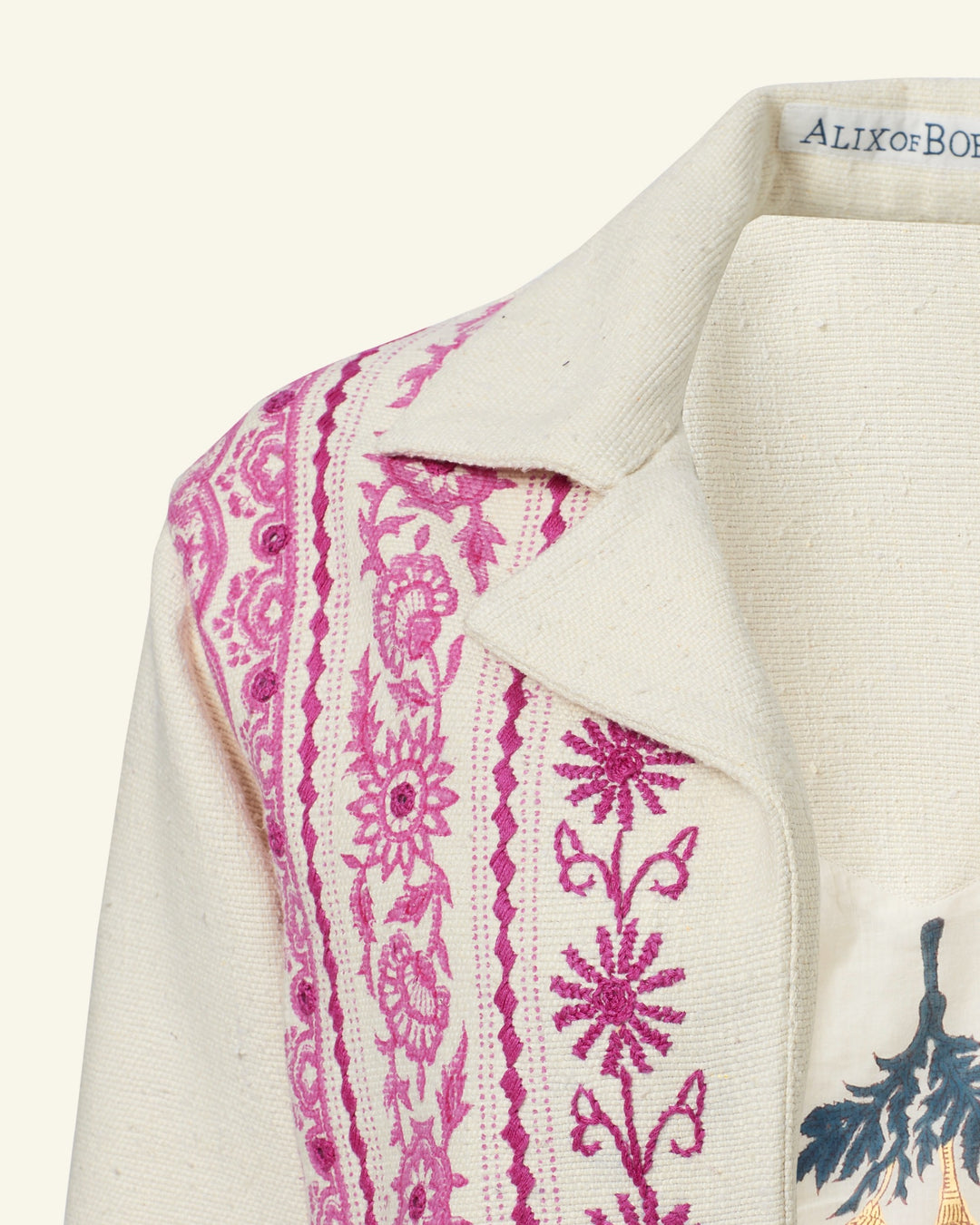 Close-up of the Western Phlox Riviera Jacket, showcasing intricate pink hand-embroidered floral and geometric patterns along the collar and shoulder on an off-white linen fabric. With a visible partial label on the inside, its breezy design makes it perfect as a summer jacket.