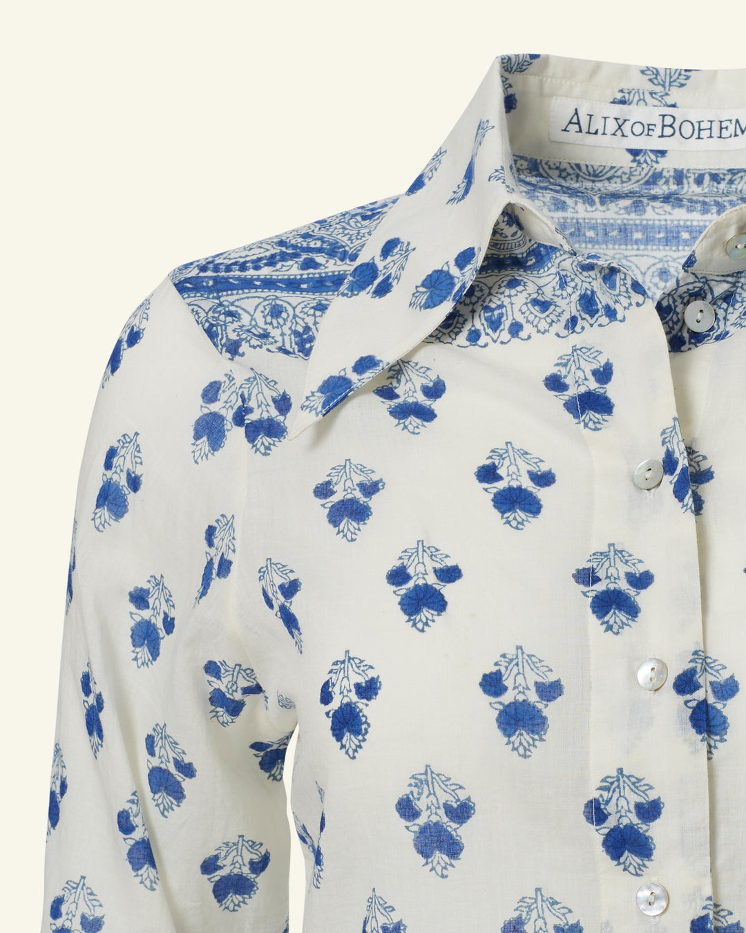 Experience an effortlessly chic look with the Dolly Indigo Bhutti Shirt, a white button-up featuring blue, hand-block printed floral and leaf patterns. The collar and shoulders are adorned with intricate blue designs. Labeled "ALIX of BOHEMIA," this shirt pairs perfectly with high-waisted pants.