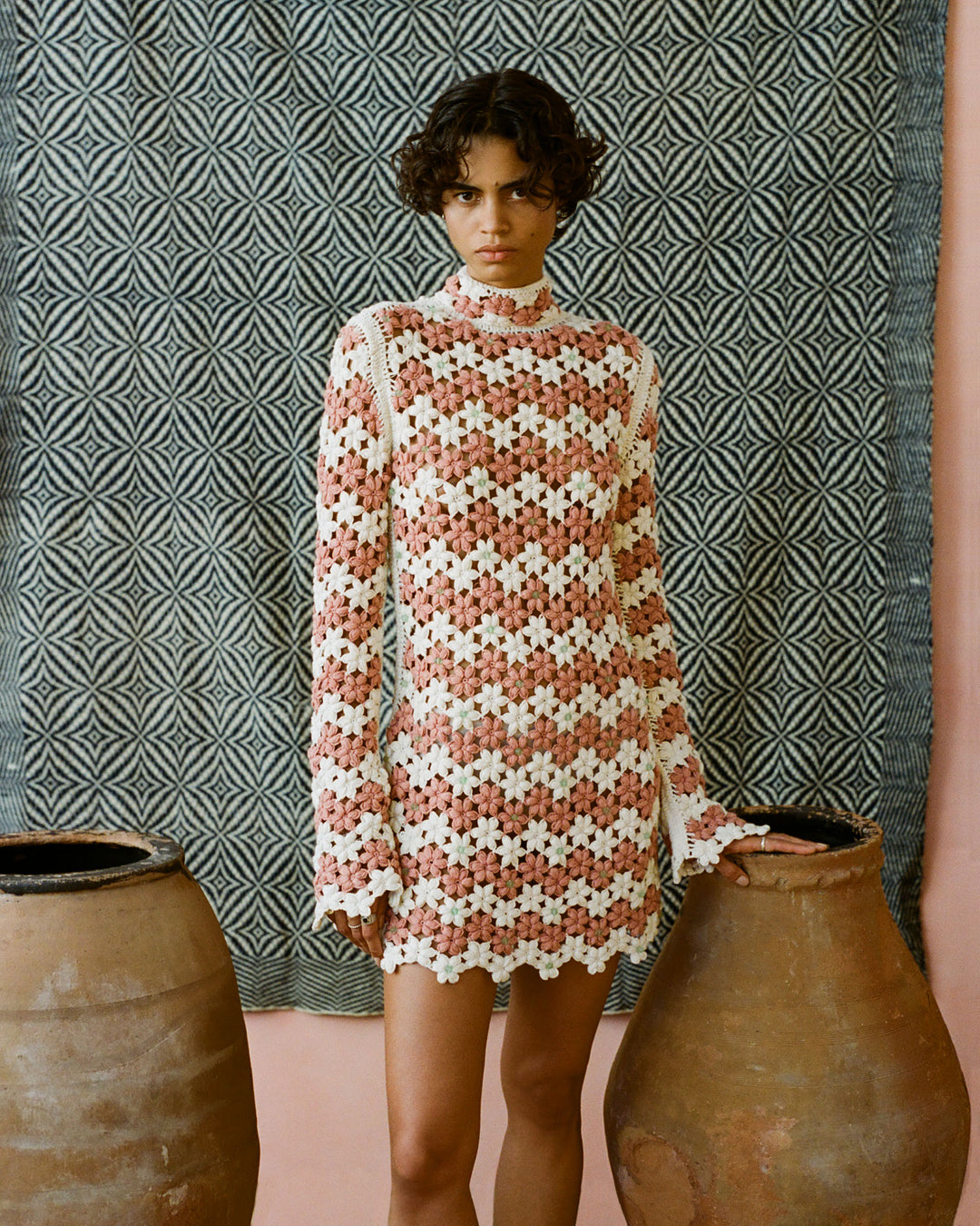 A person with curly hair stands between two large clay pots, wearing the Romy Peachy Crochet Dress, a patterned long-sleeve dress made from khadi cotton. The background features a geometric black and white woven textile.