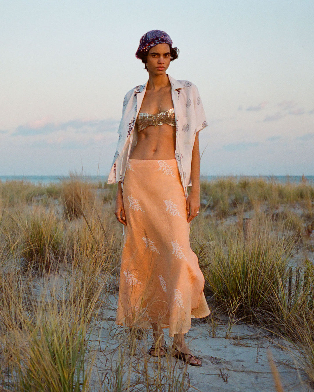 A person stands on a sandy beach with scattered grass, wearing a sequined bikini top and the Styla Porcelain Linen Shirt open over their flowing peach skirt. A purple headscarf adorns their head as the clear sky embraces a subtle sunset glow.