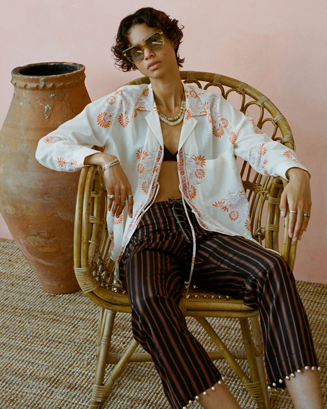 A person sitting in a wicker chair, wearing sunglasses, a white floral shirt, and Jodhpur Clove Beaded Pants. They have short hair and are accessorized with necklaces and bracelets. In the background is an earthen pot against a pink wall adorned with hand-block printed linen details.