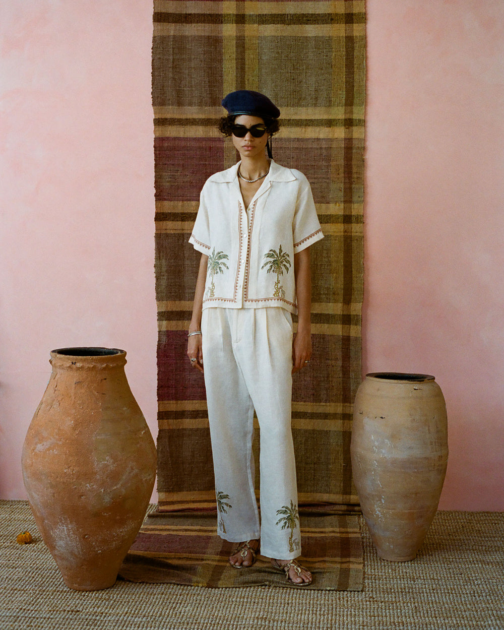 A person wearing a Stevie Sanded Palm Shirt with palm tree designs stands in front of a plaid backdrop. They accessorize with a hat, sunglasses, and sandals, surrounded by large clay pots on woven flooring.