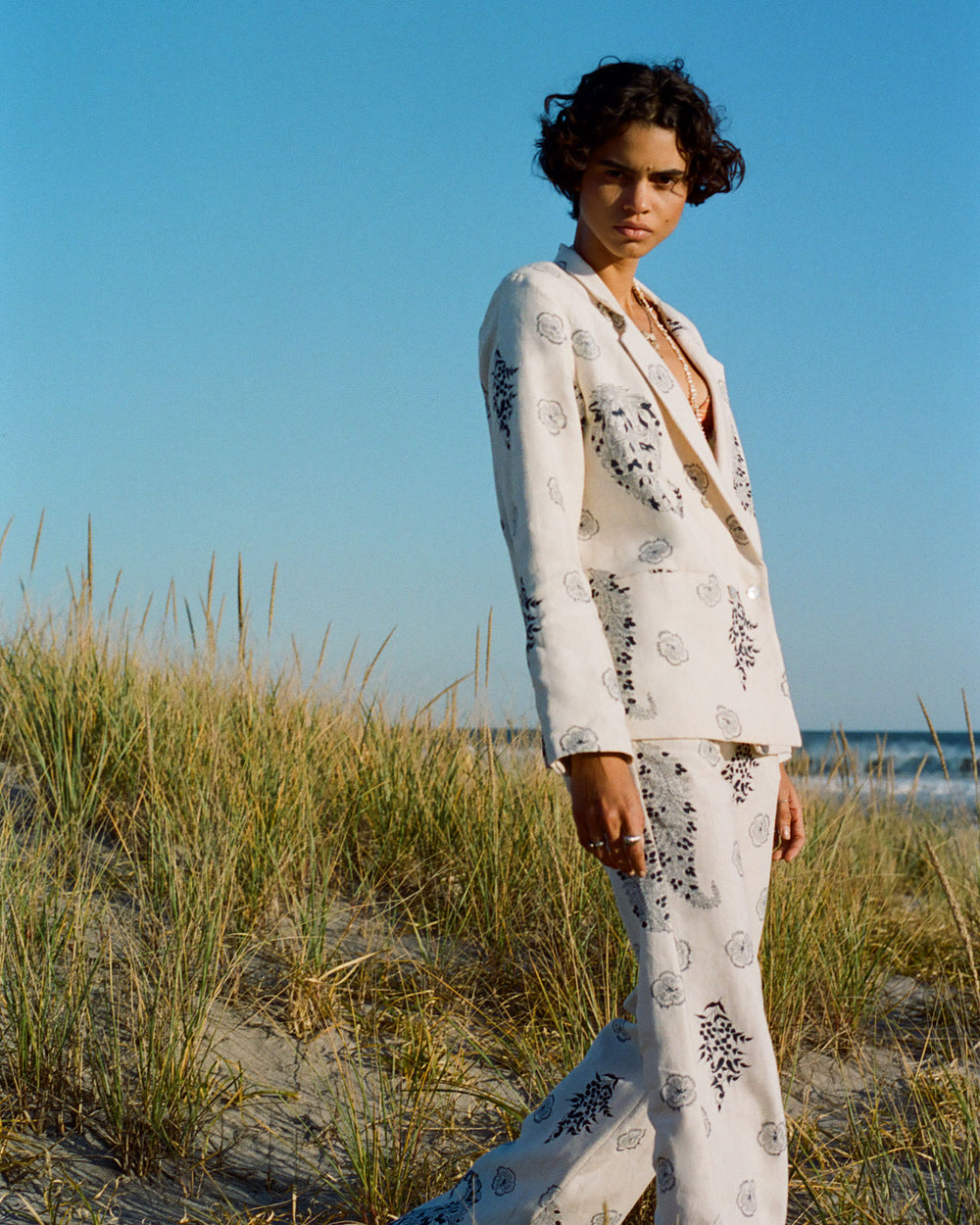 A person with short curly hair stands outdoors on a sandy beach, wearing a white suit with black patterns. The Charlie Porcelain Linen Pant, featuring subtle floral motifs, adds charm to the ensemble. Tall grass is visible in the background under a clear blue sky.