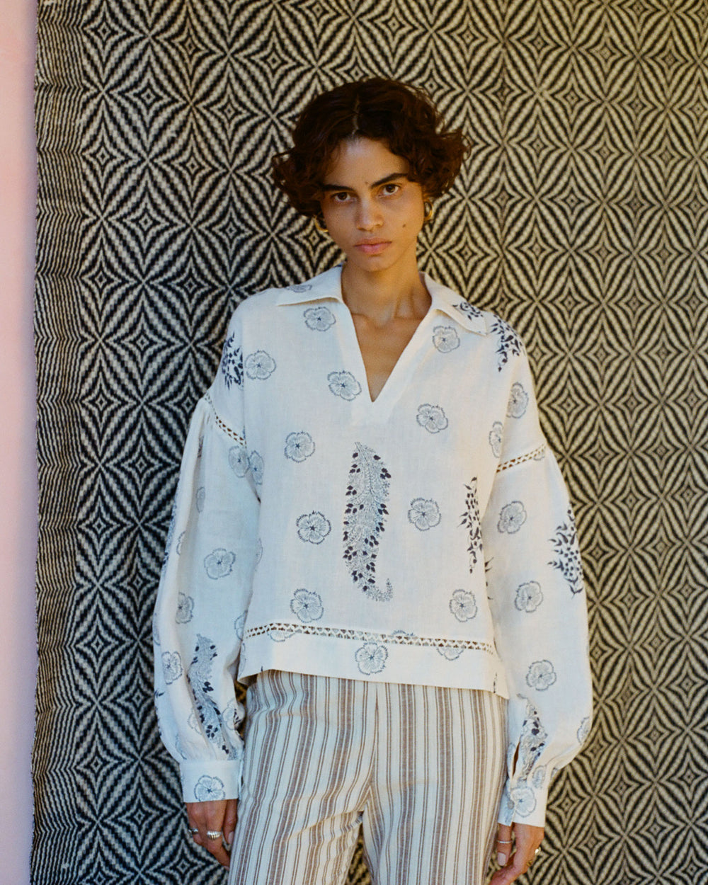 A person with short curly hair stands in front of a patterned black and white backdrop, wearing the Dylan Porcelain Linen Top—a loose, embroidered white blouse with floral designs. This is complemented by pinstripe pants and an elegant hand-block printed floral print blazer.