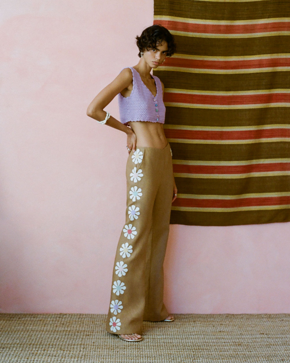 A person stands against a pink wall with a striped tapestry, wearing the Ludi Lavender Crochet Top and brown wide-leg pants adorned with white and red flower designs. The floor is covered with woven material.