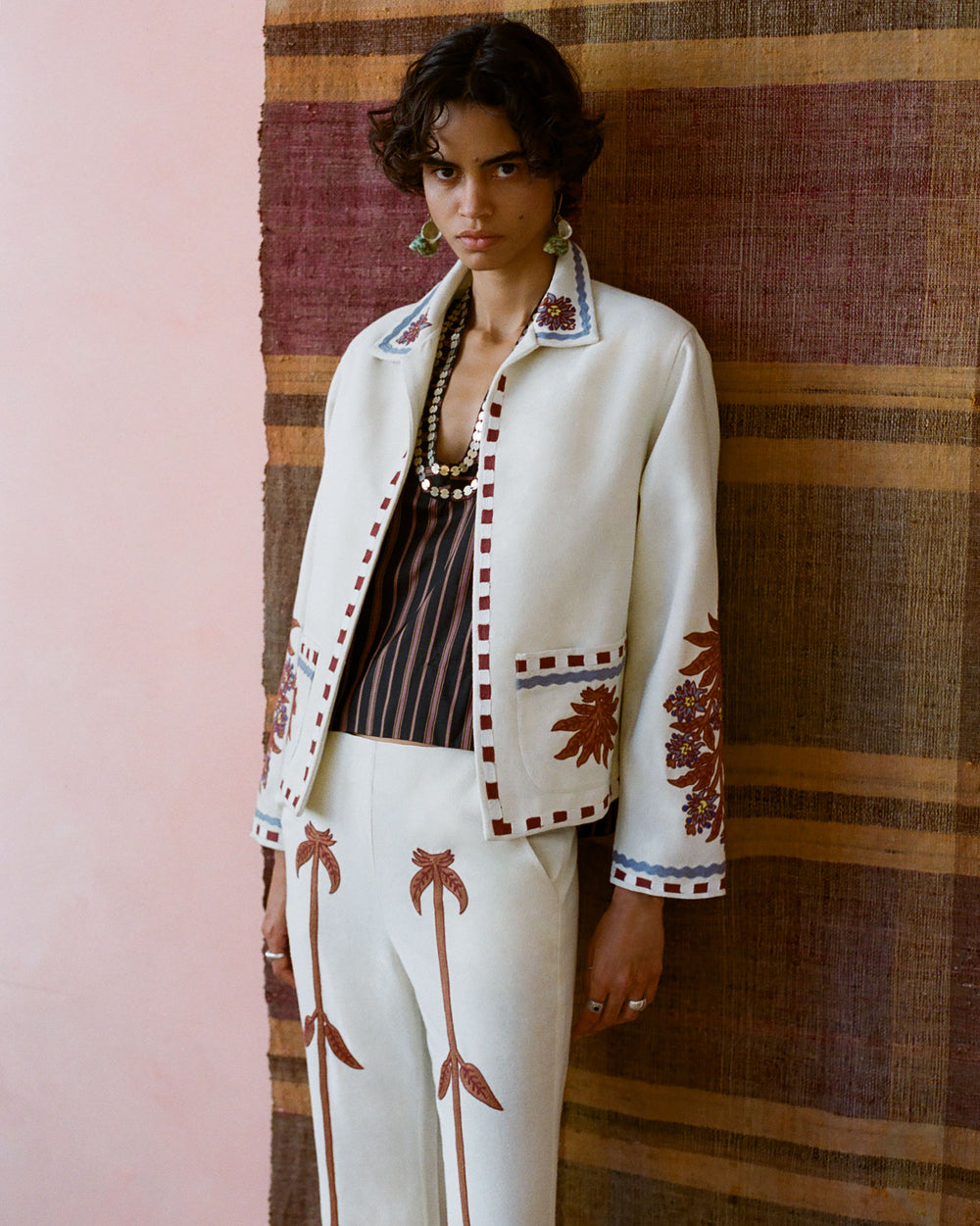 Introducing someone dressed in the Francis Waterlily Bloom Jacket, a cream-colored piece featuring hand-embroidered floral details, standing against a textured, earth-toned backdrop. The ensemble includes matching pants embellished with striking red flower and leaf motifs, complemented by short, wavy hair and statement earrings.