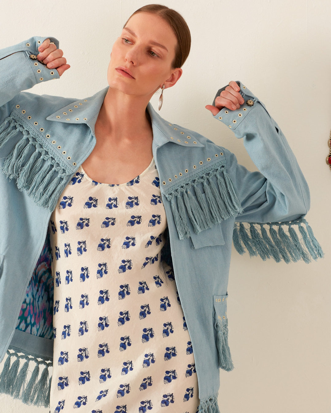 A person wearing the Jude Aqua Fringe Jacket and a white dress with blue patterns stands against a plain background. The jacket is pulled slightly off their shoulders, showcasing the beauty of its hand-embellished details. Their hair is neatly tied back, and they wear a single earring.