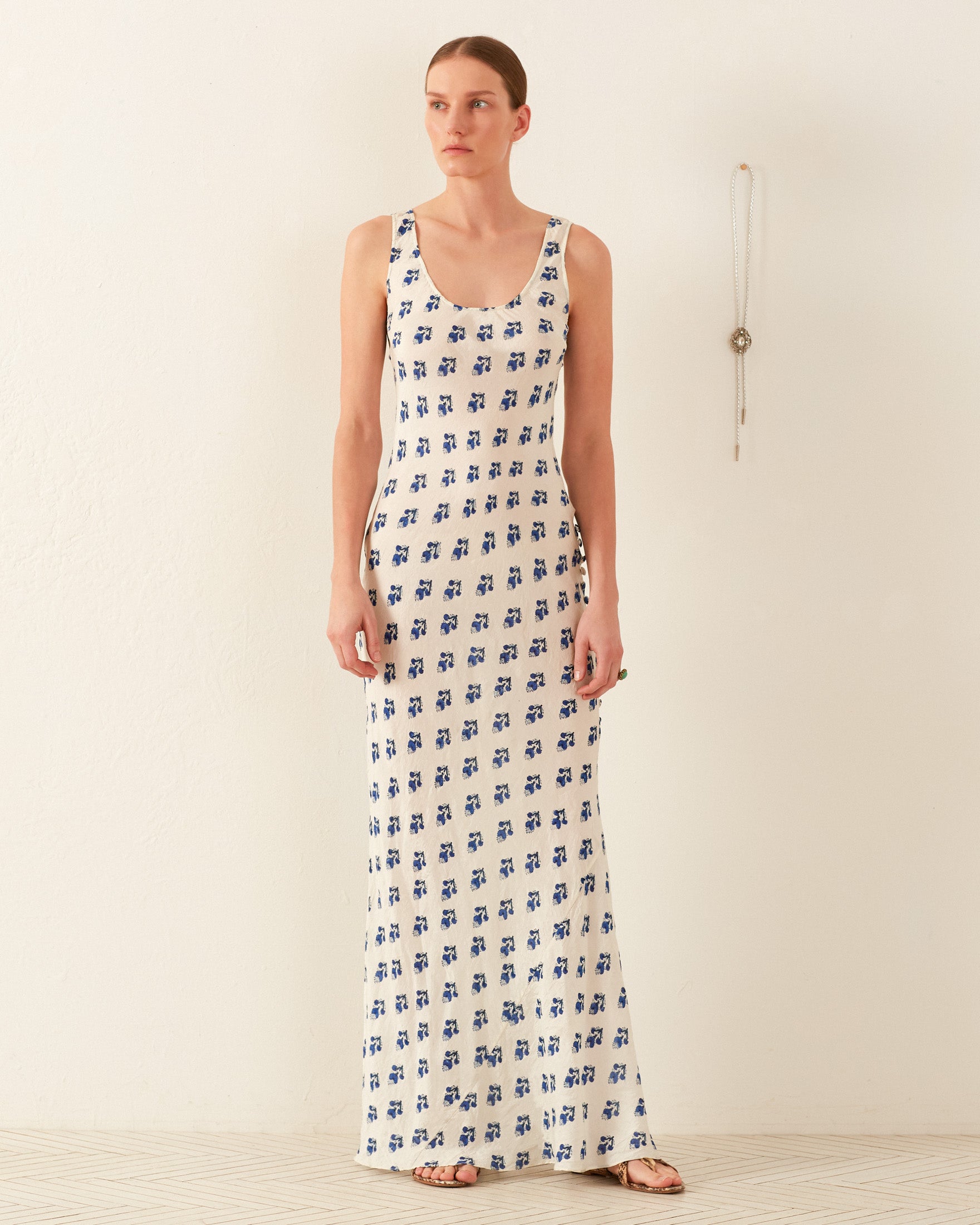 Wearing the Adele Indigo Bhutti Silk Dress, a person stands against a plain white wall, showcasing its sleeveless, floor-length hand-block printed design with a blue floral pattern and '90s-inspired silhouette. A necklace hangs on the wall beside them, beautifully complementing their sandals and ring.