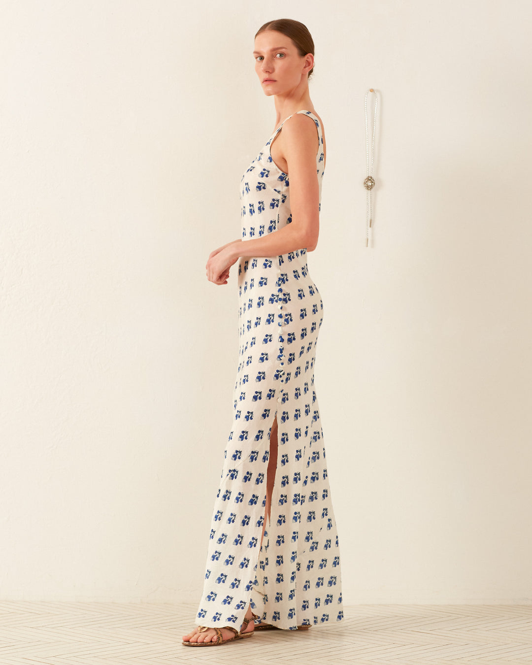 A person wearing the Adele Indigo Bhutti Silk Dress, a '90s-inspired white silk slip dress adorned with blue floral patterns, stands in profile against a plain background. A necklace with a pendant hangs on the wall.