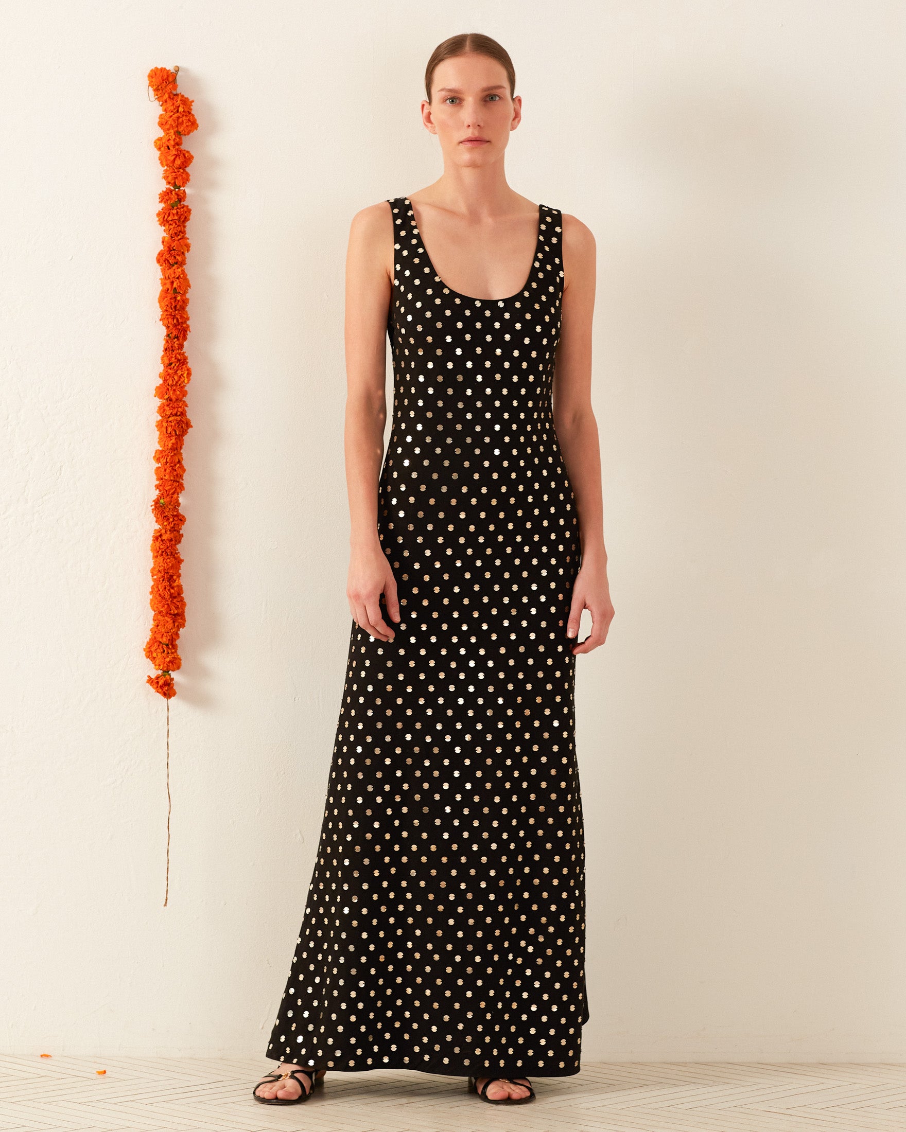 A person stands wearing the Adele Night Sky Coin Dress, a sleek black artisan garment adorned with white polka dots and sleeveless design, paired with black sandals. A vibrant vertical garland of orange flowers adorns the wall beside them, enhancing the elegance of the plain light-colored backdrop.