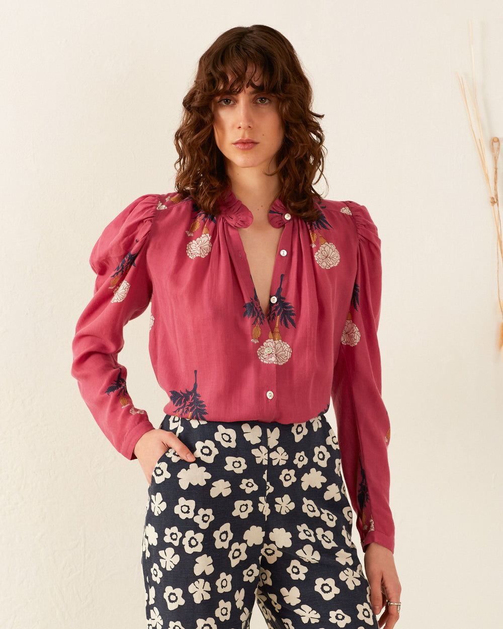 A person with wavy brown hair wears the Arte Hibiscus Rose Shirt, a pink blouse featuring floral details, paired with black and white floral patterned pants. A cropped blazer adds a vintage feel as they stand against a neutral background with a minimalistic design.