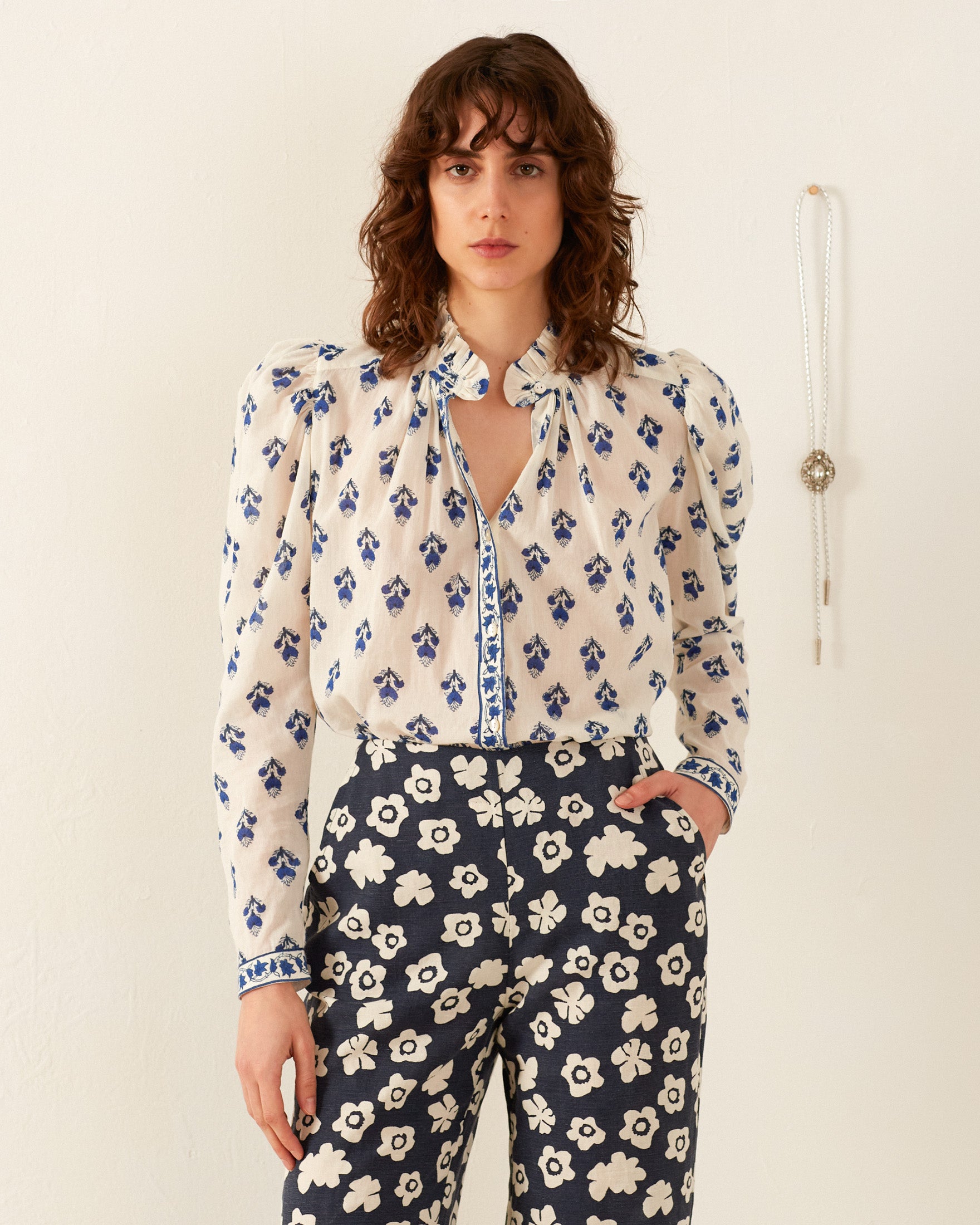 A person stands against a white wall, wearing the Artemis Indigo Bhutti Shirt, which features hand-block printing, puffed sleeves, and a tie-neck detail. They pair it with trousers showcasing a pink floral pattern. A long necklace with a pendant is hanging on the wall nearby.