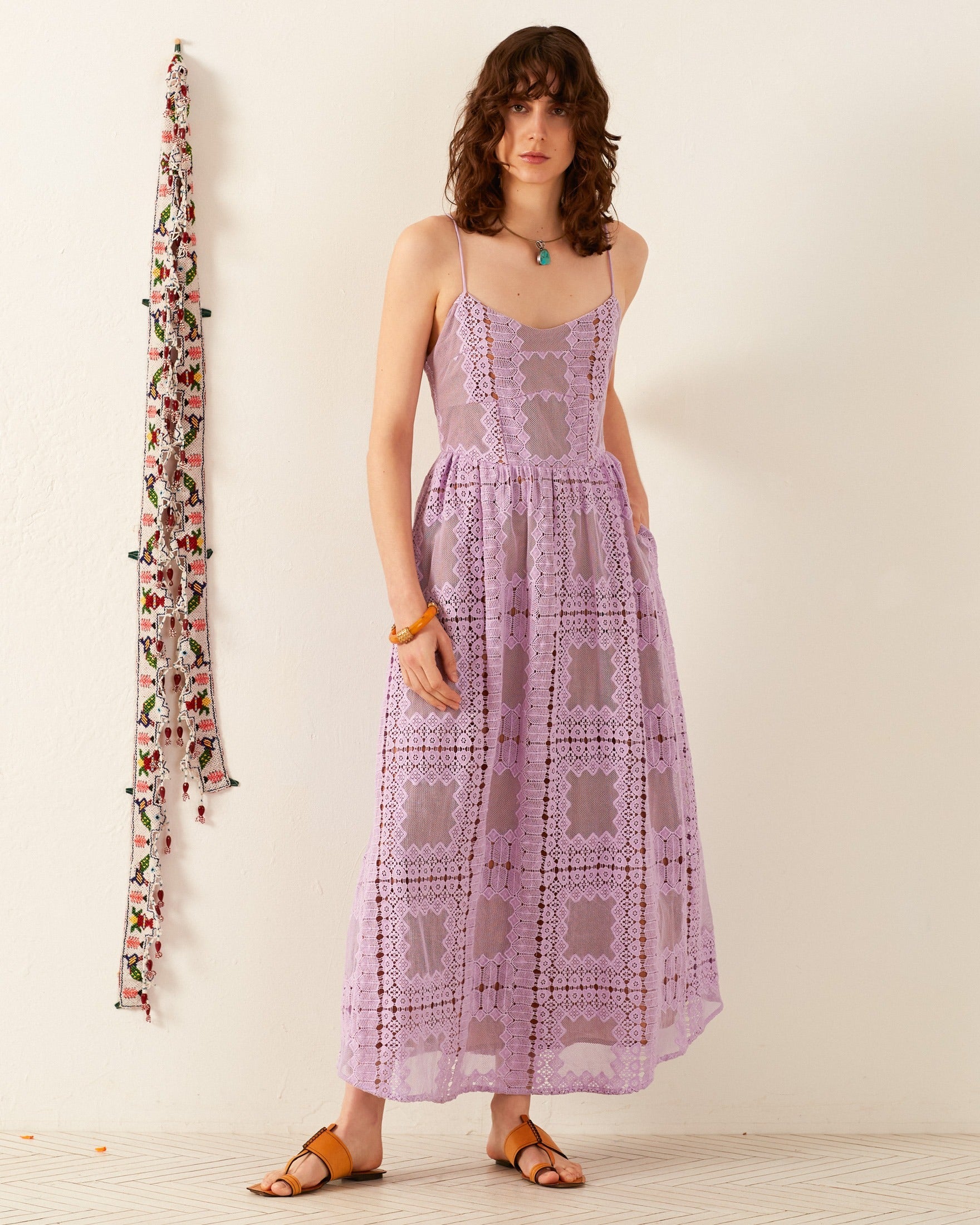 A woman stands against a white wall, elegantly dressed in the Autumn Lavender Picnic Dress featuring delicate lace and thin straps. Her curly brown hair complements her turquoise necklace and wooden bracelet. She completes her wedding guest attire with brown sandals. A colorful, floral wall decoration is on her left.