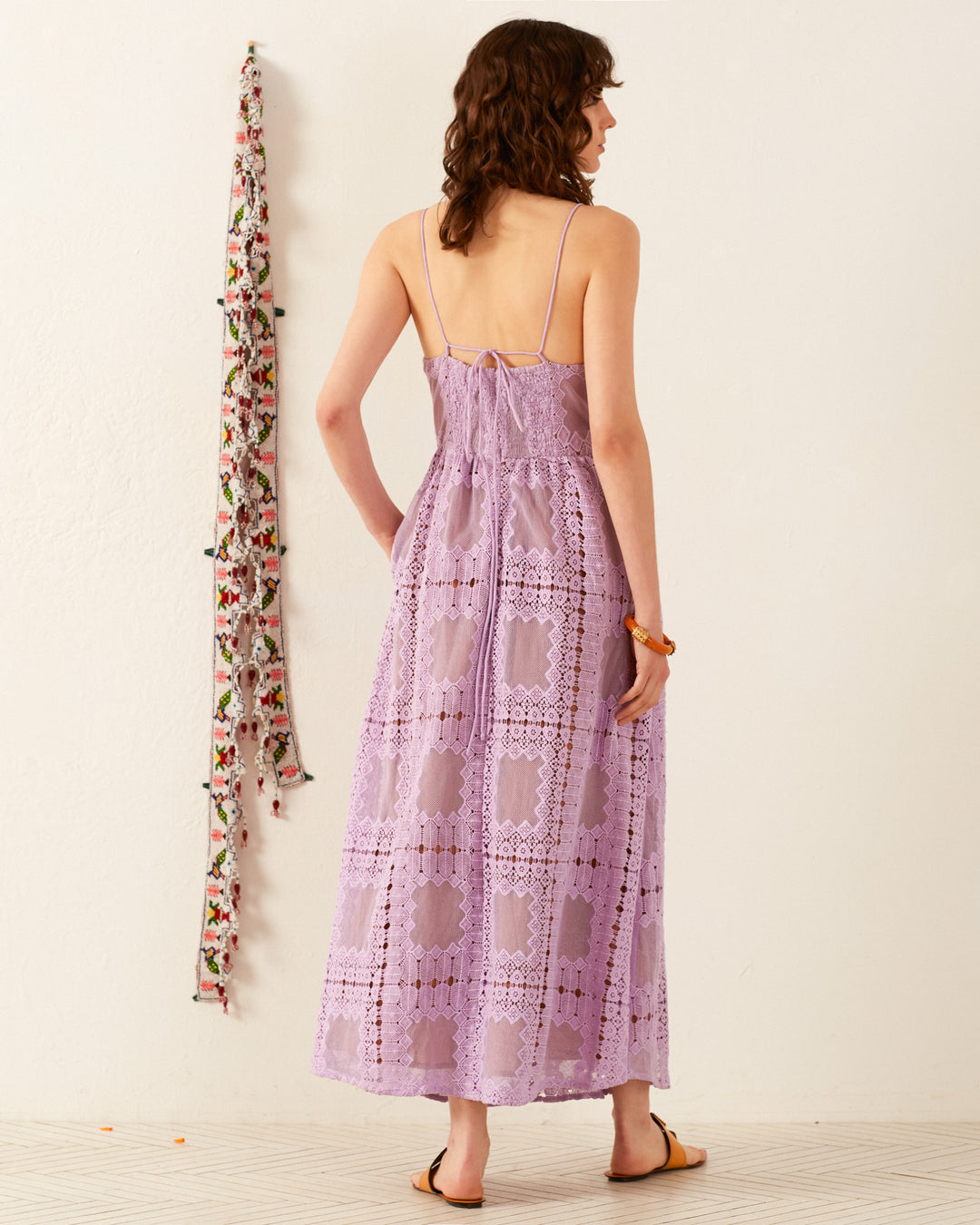 A person with wavy brown hair is wearing the Autumn Lavender Picnic Dress, a sleeveless crochet midi dress, along with sandals, perfectly suited for wedding guest attire. They're standing in a room adorned with a decorative floral wall hanging.