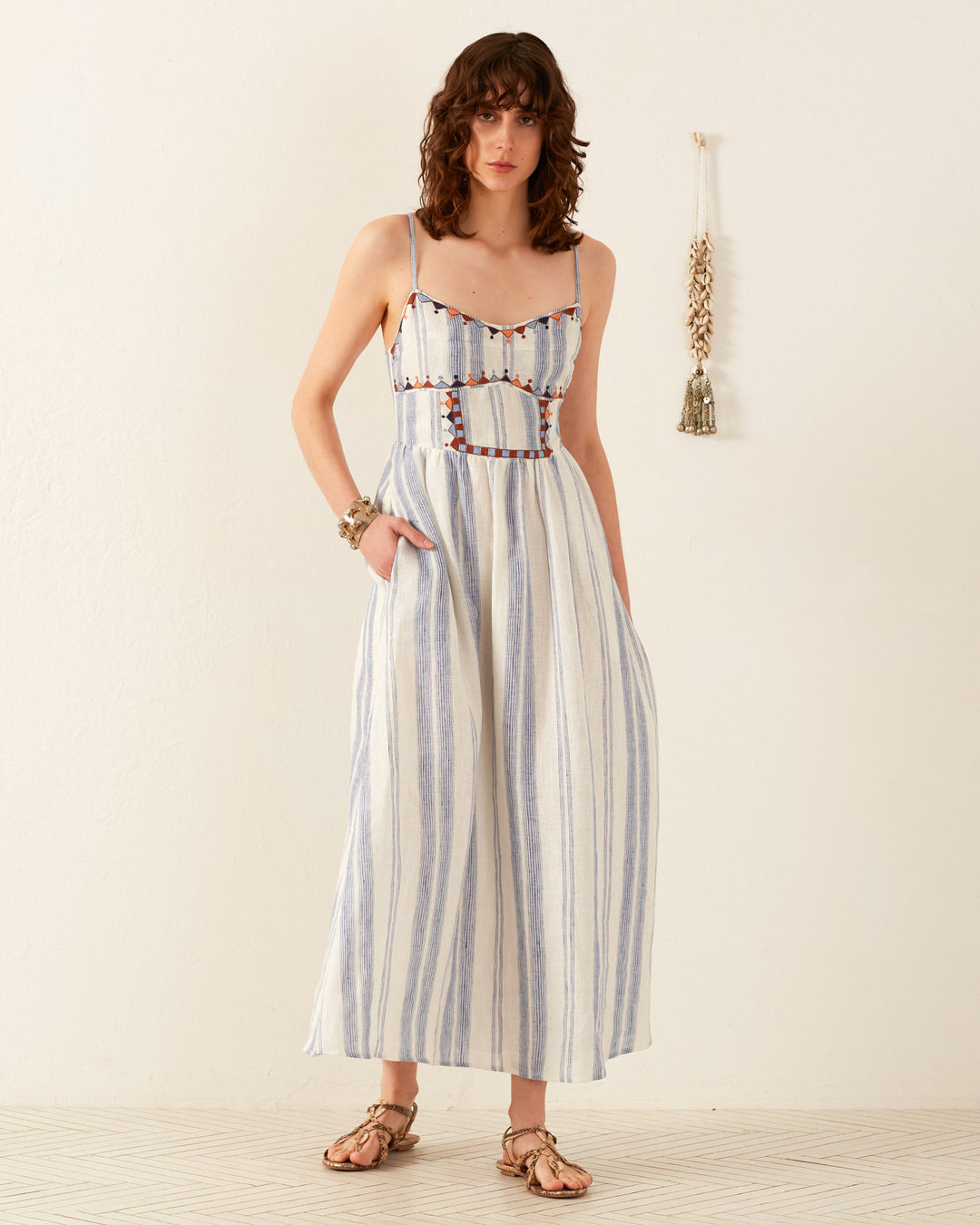 A woman wearing the Autumn Pacific Delta Dress, a sleeveless midi dress with blue and white stripes and embroidered details, poses against a plain light-colored background. Perfect for summer occasions, she stands with her hands in the dress pockets, complementing the look with sandals and a bracelet.