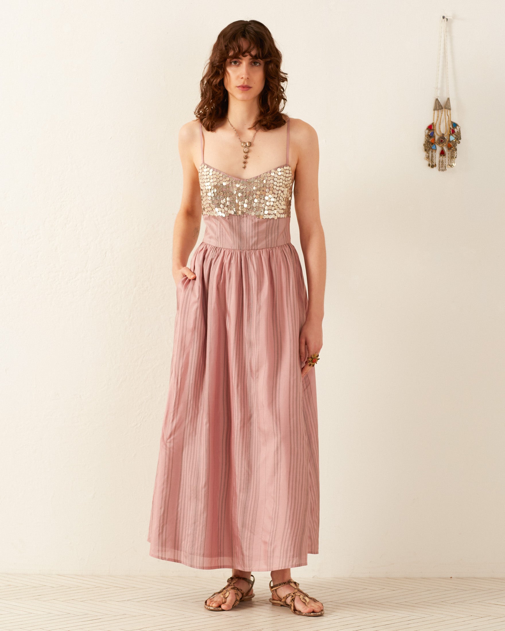 Against a white wall, a person showcases the Autumn Petal Coin Dress, a pink, floor-length gown highlighted by a sequined bodice and delicate thin straps. Ideal for summer occasions, they enhance their ensemble with sandals as a decorative wall hanging lends additional charm to the scene.