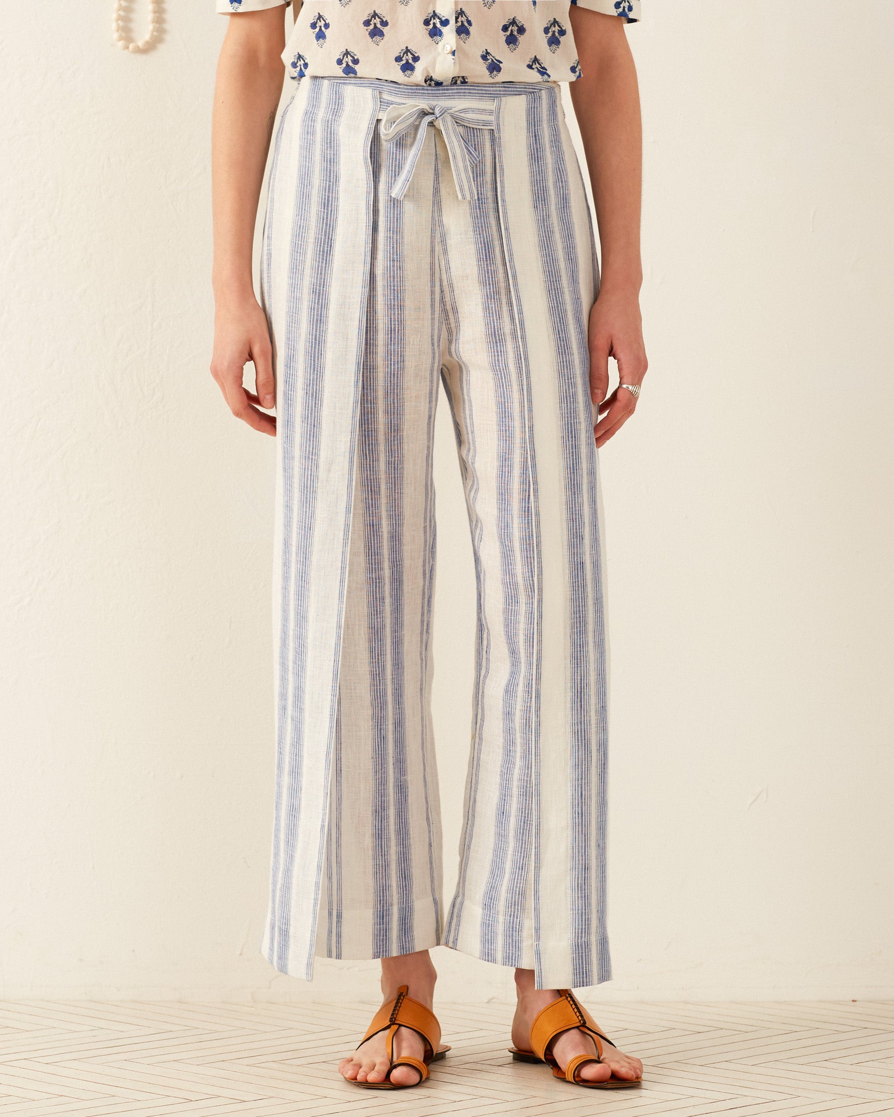 A person is wearing the Bonnie Pacific Wrap Pant, which features light blue and white vertically striped fabric tied with a front bow, reminiscent of a vintage-inspired dress. They pair it with a white top adorned in blue patterns and brown sandals, set against a neutral light background.