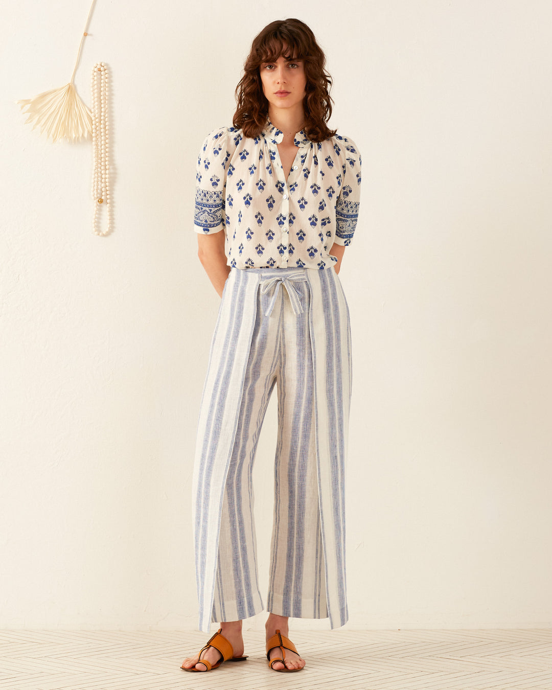 A person wearing a white blouse with blue patterns and the Bonnie Pacific Wrap Pant stands against a plain wall. Their neutral expression complements the silk chiffon elegance in their style. Sandals complete the look, while a wall decoration hangs to the left.