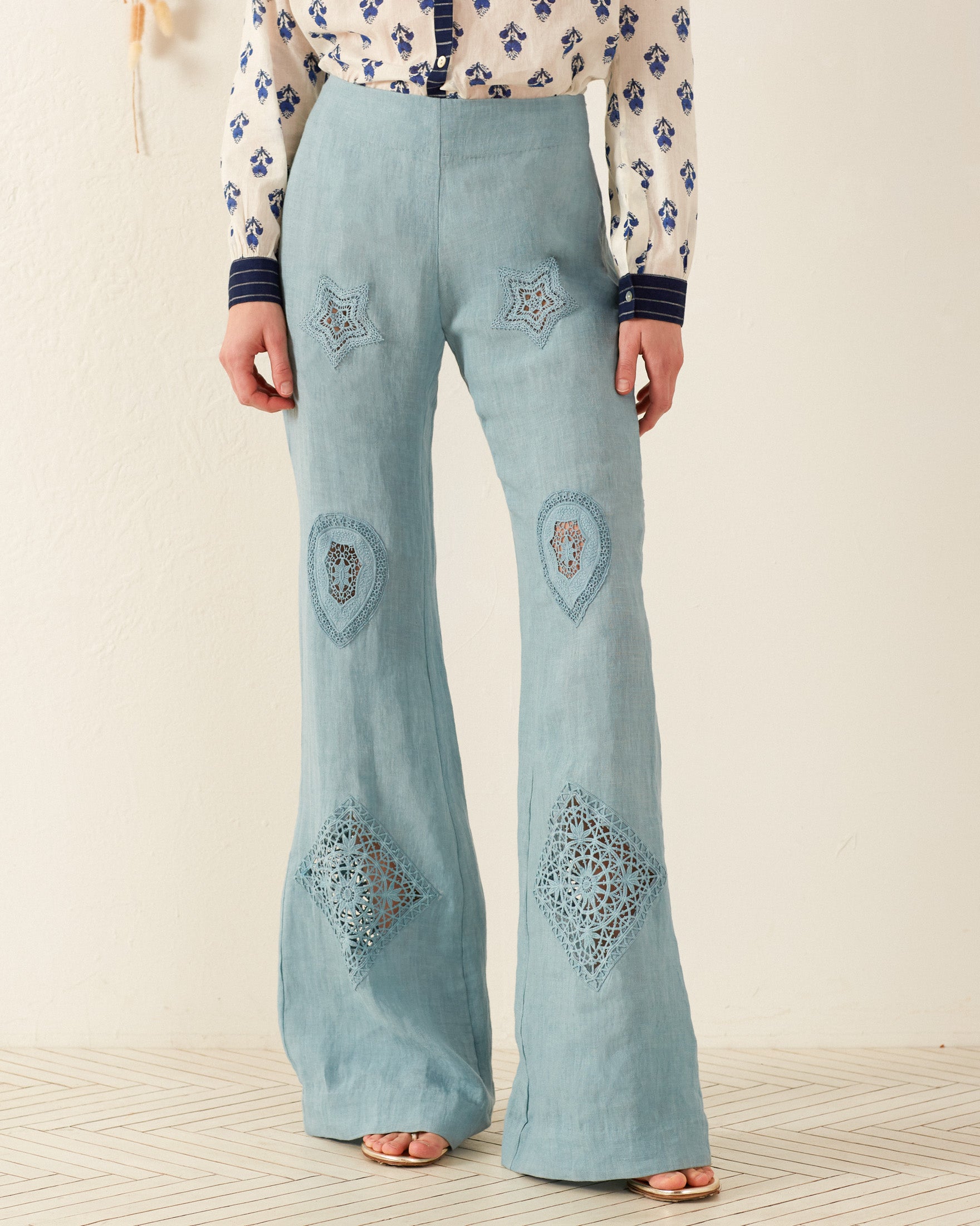 Wearing the Charlie Aqua Eyelet Pant, which features intricate star and diamond-shaped lace patterns in light blue, a person exudes a vintage-inspired feel. The pants are paired with a white blouse adorned with blue floral designs, and tan open-toed sandals complete the look.