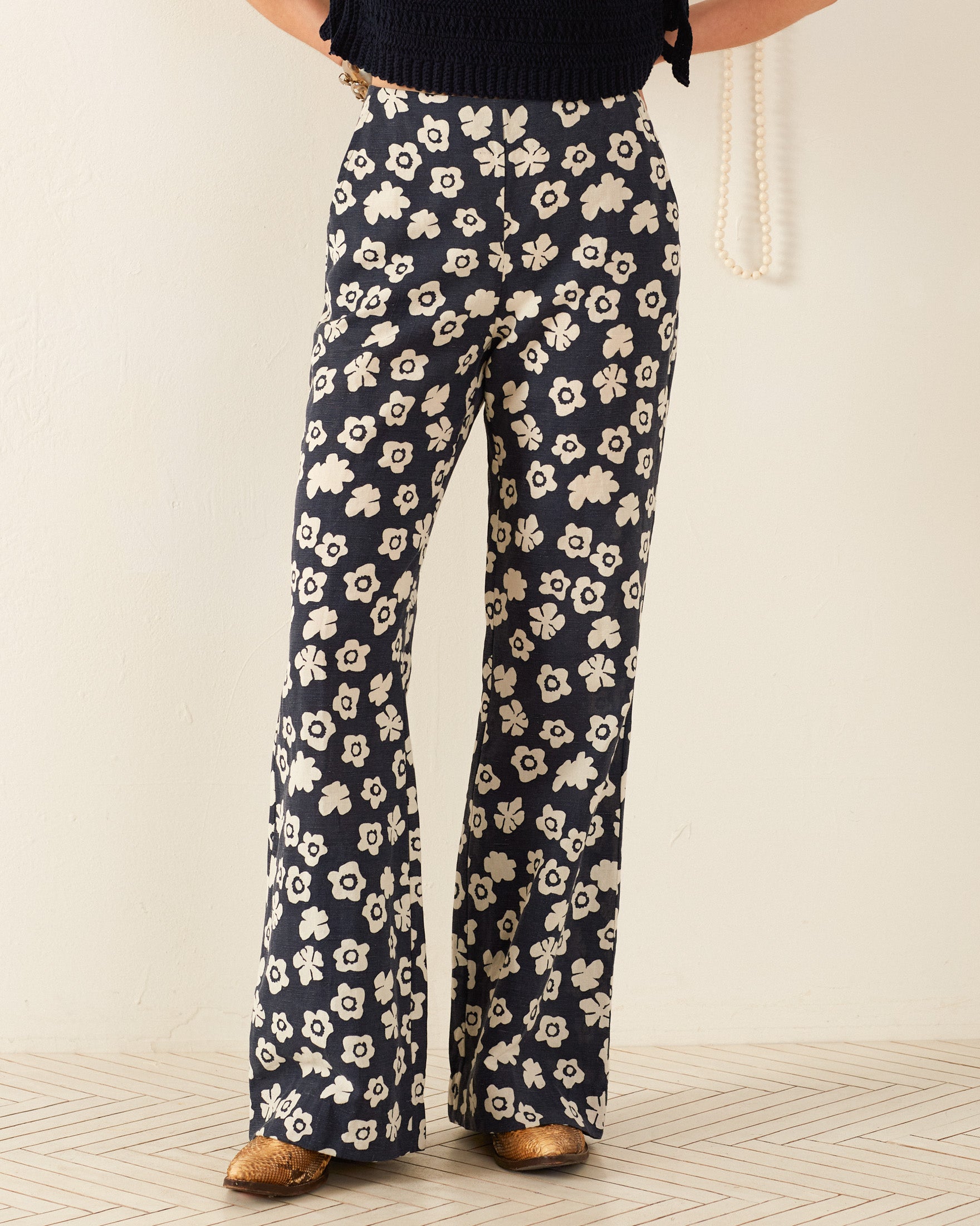 A person wearing the Charlie Deep Sea Lotus Pant, featuring a '70s-style wide-leg design with a white floral pattern against a navy background, stands against a light backdrop. The outfit includes a black top and brown shoes, beautifully highlighting the pants' retro design.